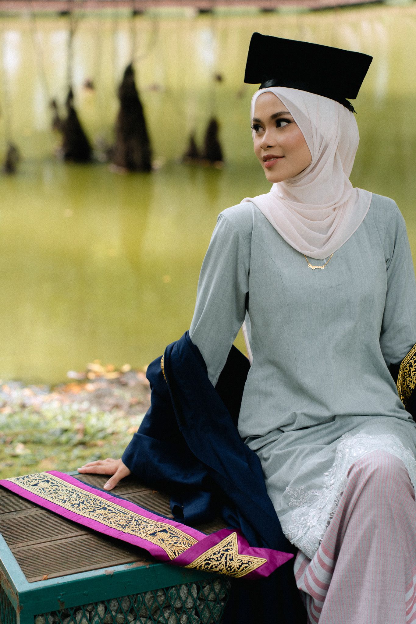 A young Muslim woman in hijab and graduation cap sits gracefully by a serene lake, wearing a elegant gray baju kurung.