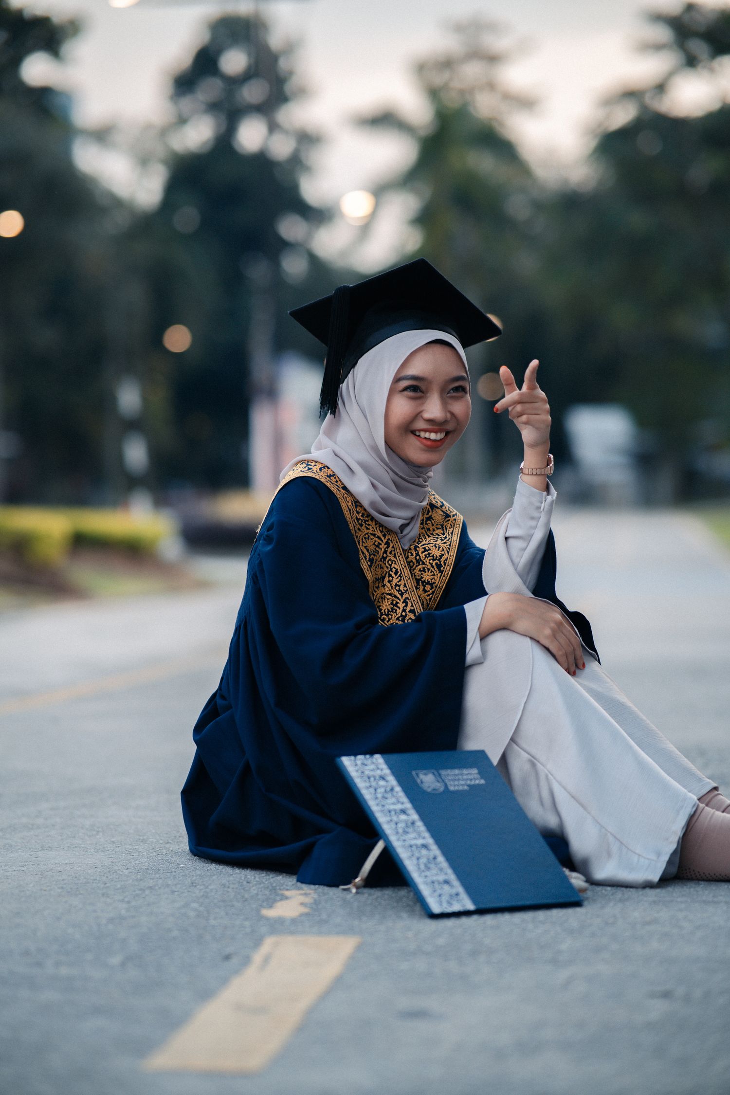 Graduate in decorated blue gown sitting on ground making peace sign.
