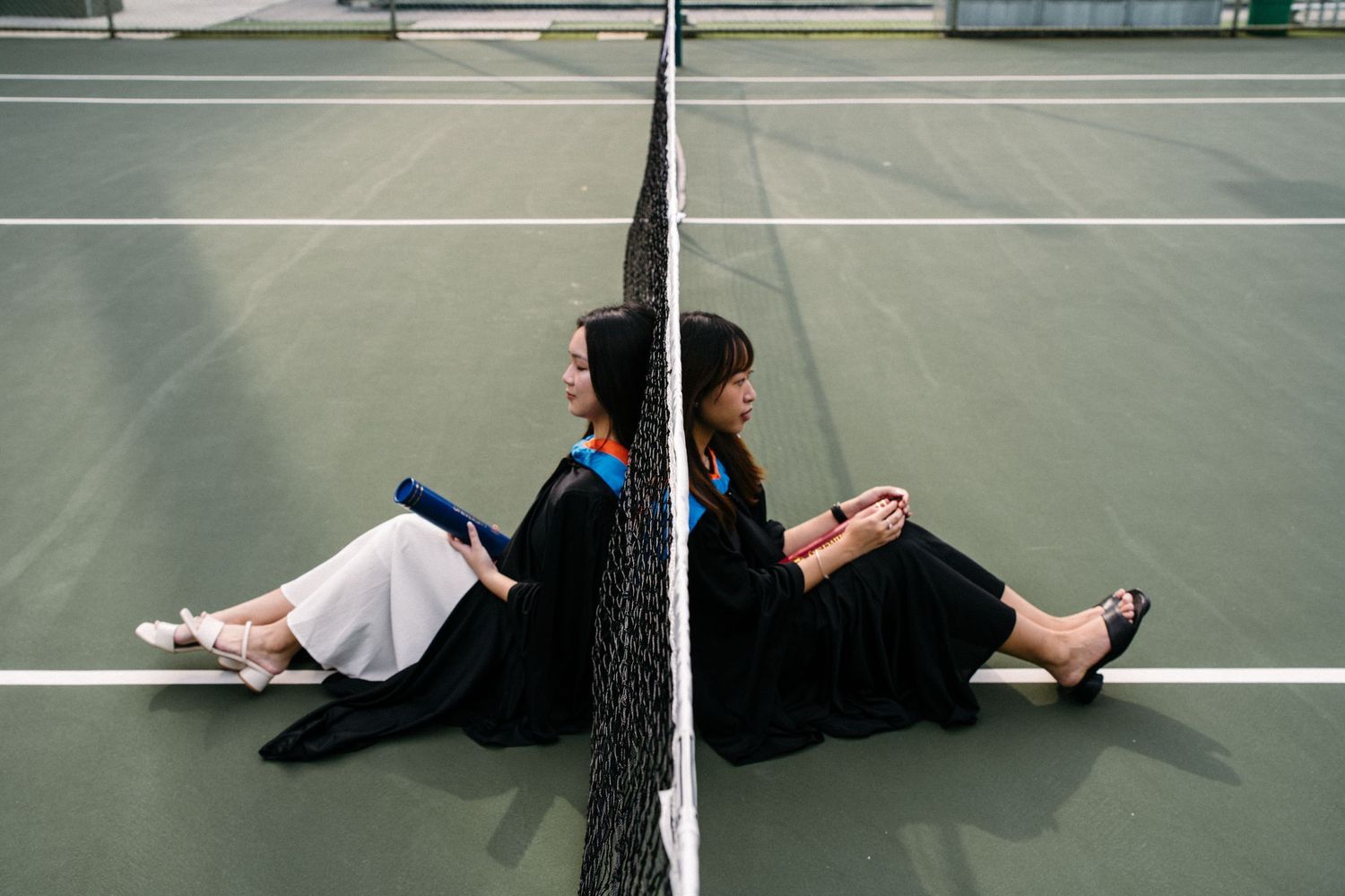 Graduate in academic attire sitting on tennis court against net reflection.