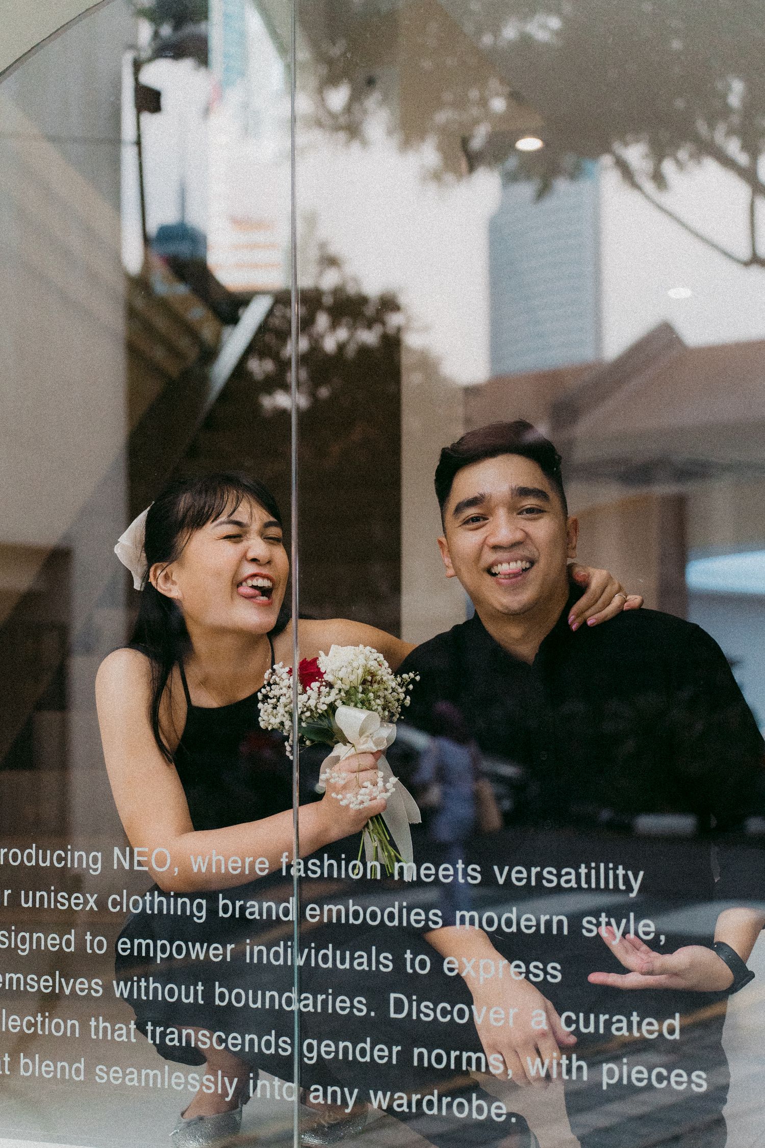 A joyful couple posing with flowers in front of a storefront window with reflective text.