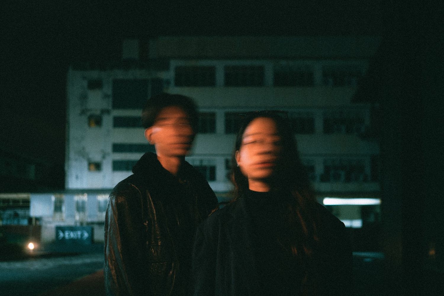 Motion-blurred portrait of two people at night with an urban building in the background.