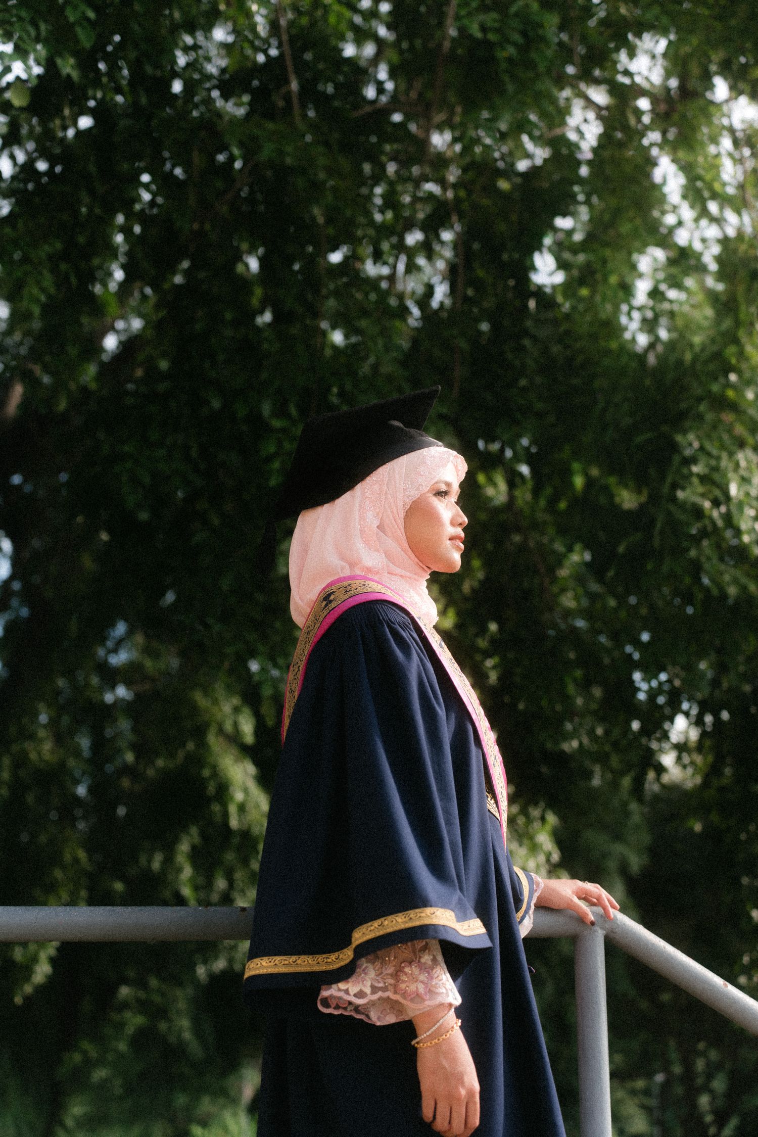 A person in a navy blue outfit and pink headscarf stands thoughtfully against a backdrop of green trees.