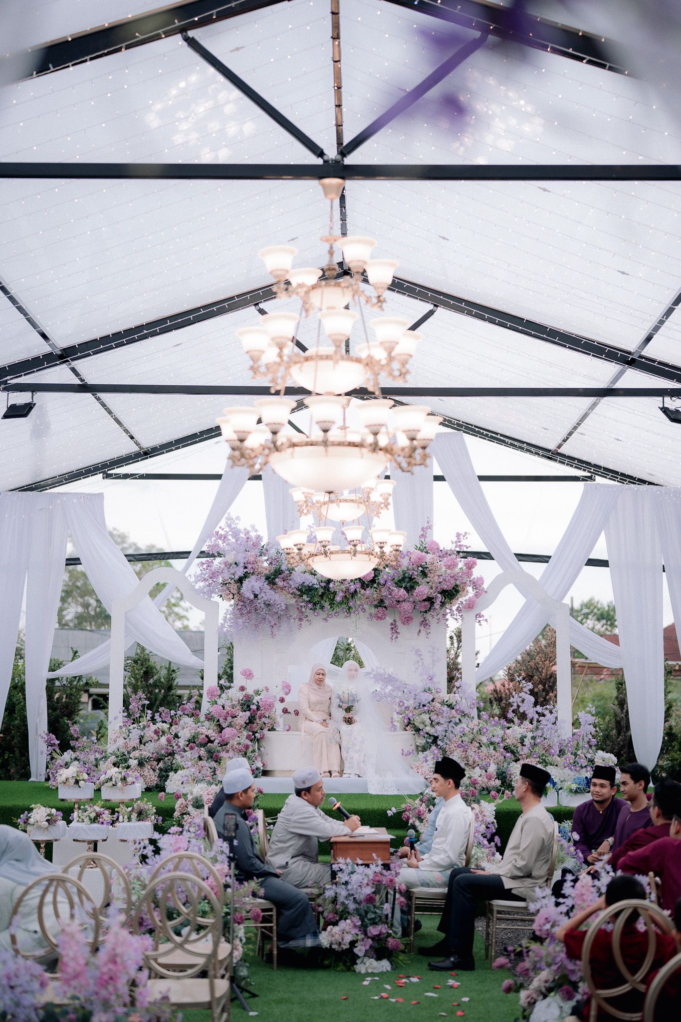 Elegant outdoor wedding ceremony under a glass tent with chandelier, purple floral decor, and guests seated on green lawn.