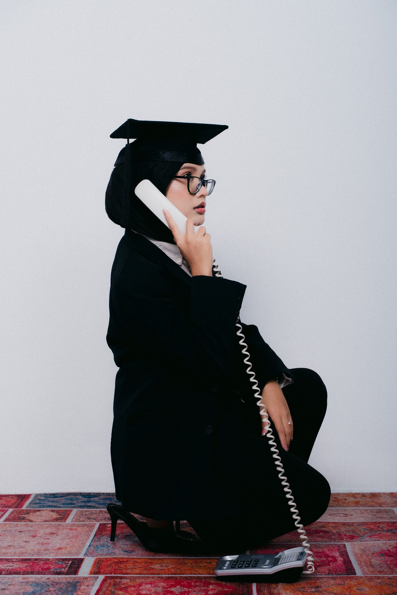 Graduate in black cap and gown sitting on floor, wearing glasses, talking on corded telephone handset.