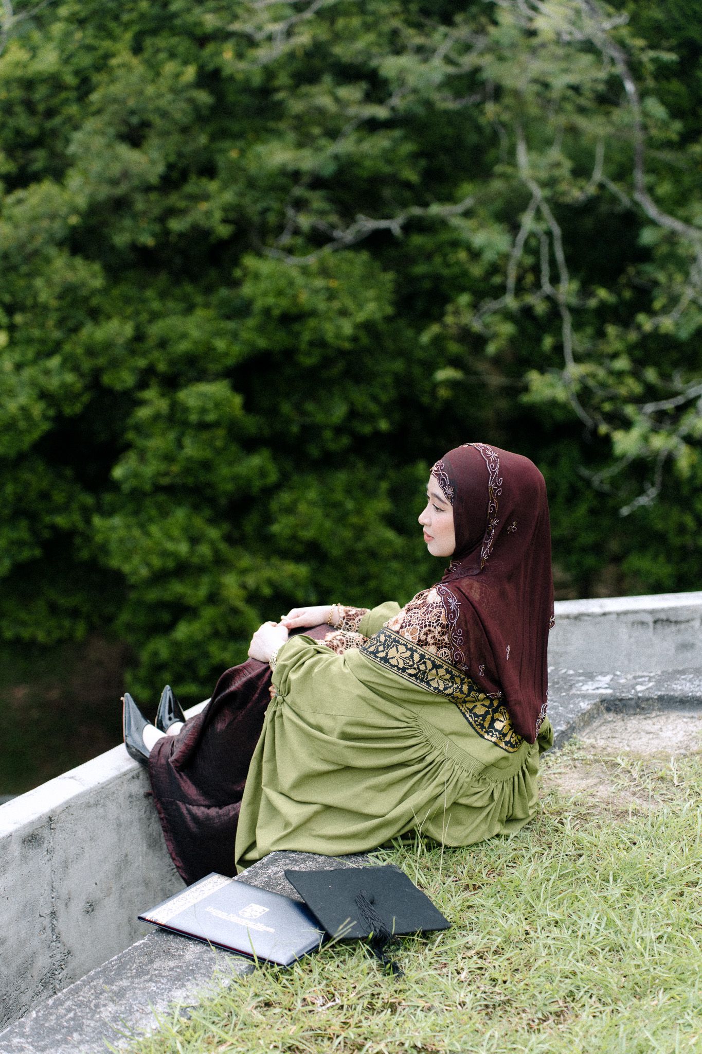 Muslim woman in hijab and green dress sitting outdoors against a wall, relaxing near an open book with lush trees behind.