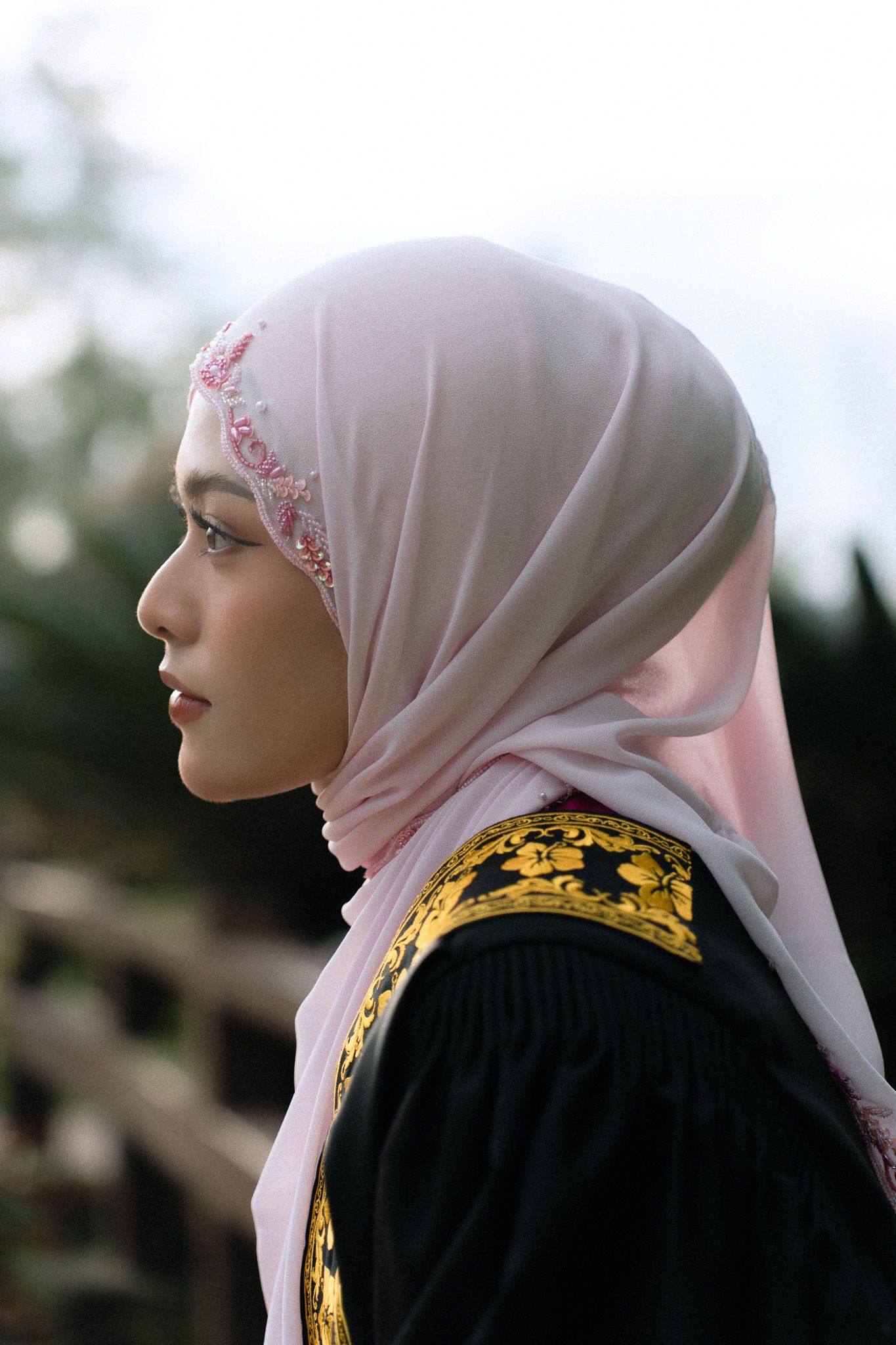 Young woman in profile wearing a pink embroidered hijab and black traditional dress with gold detailing outdoors.