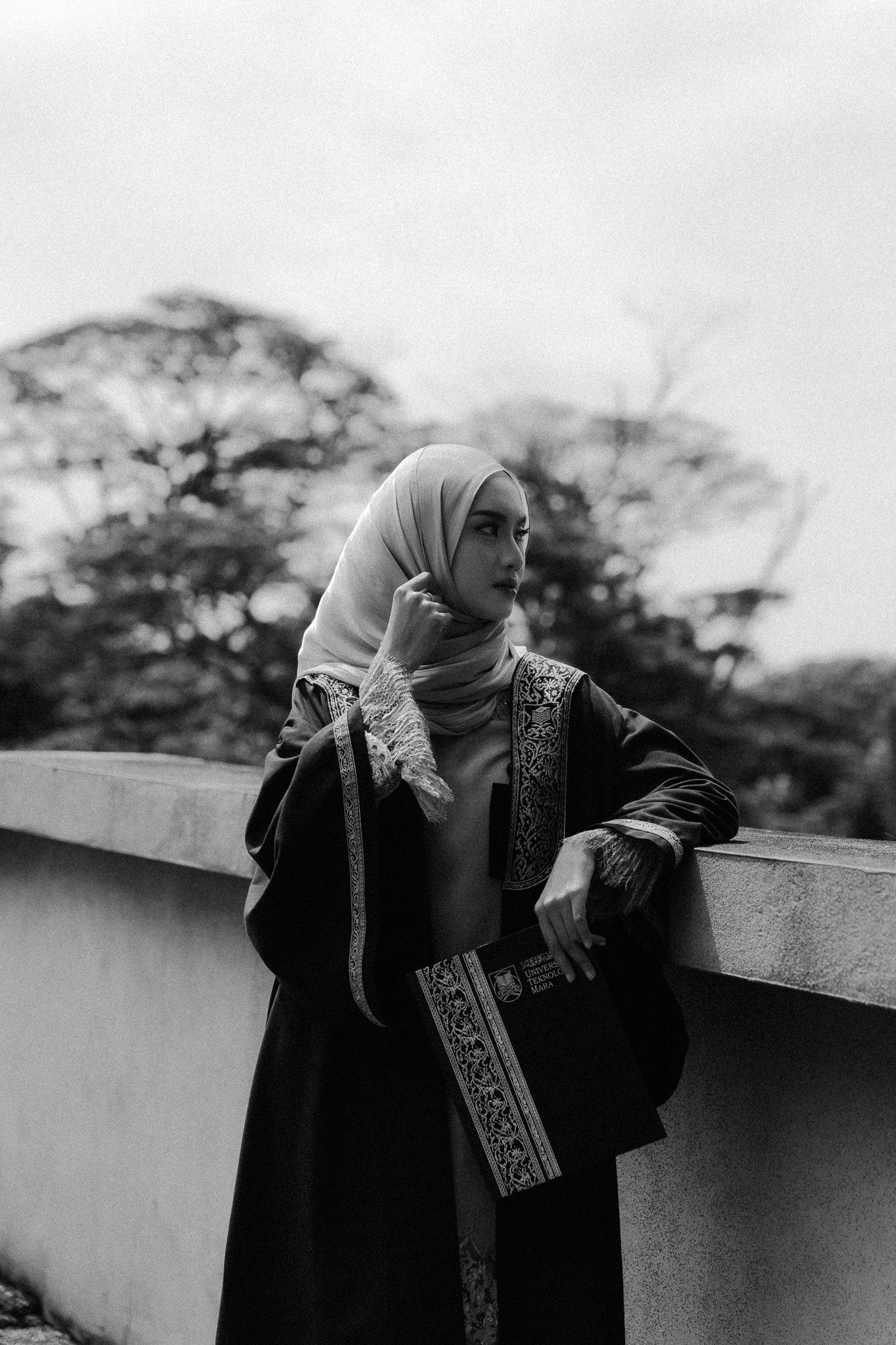 Black and white portrait of a woman in hijab and embroidered abaya leaning against a wall with trees in background.