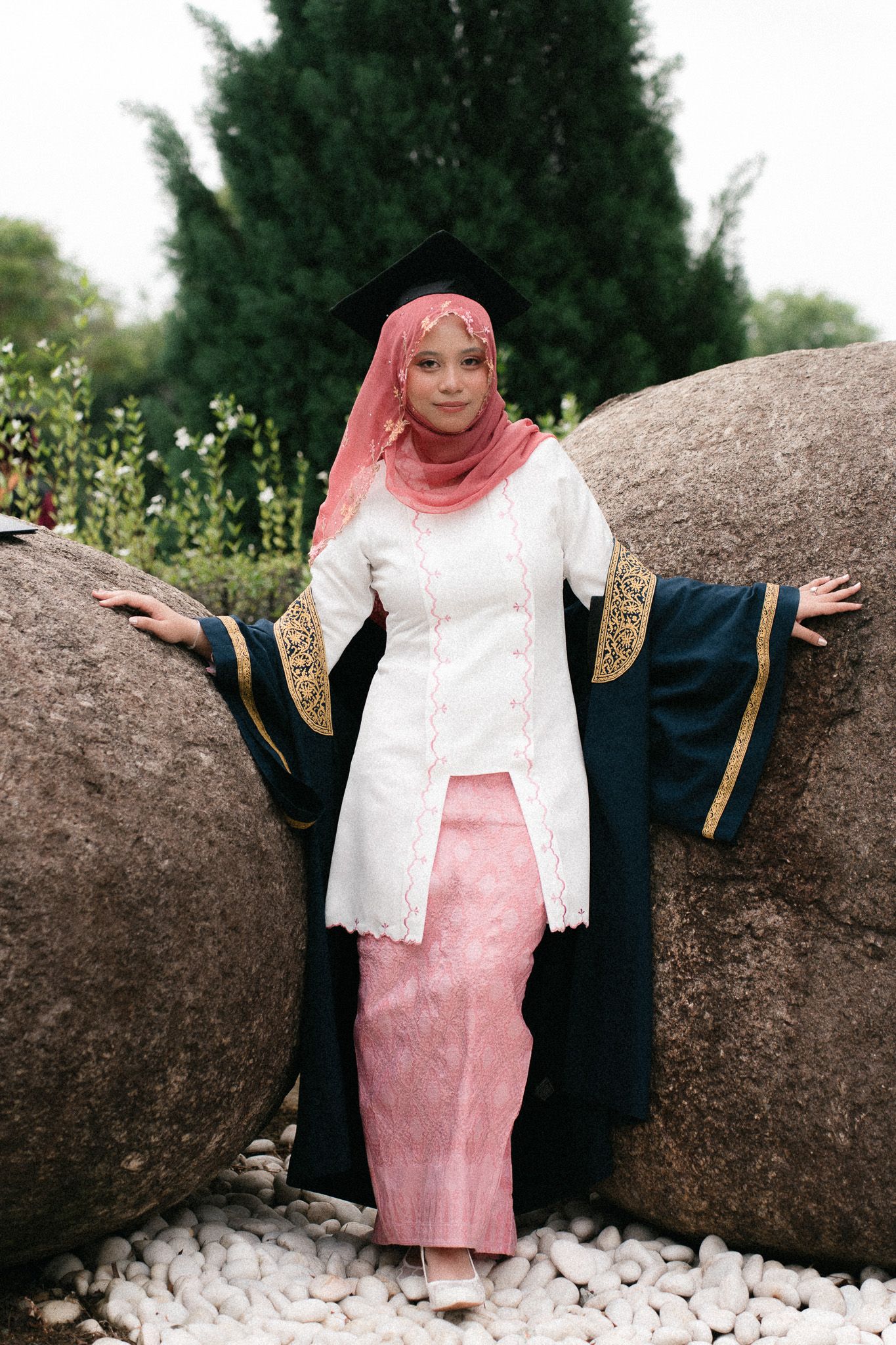 Muslim woman in graduation cap and gown with pink hijab posing between large rocks in a garden.