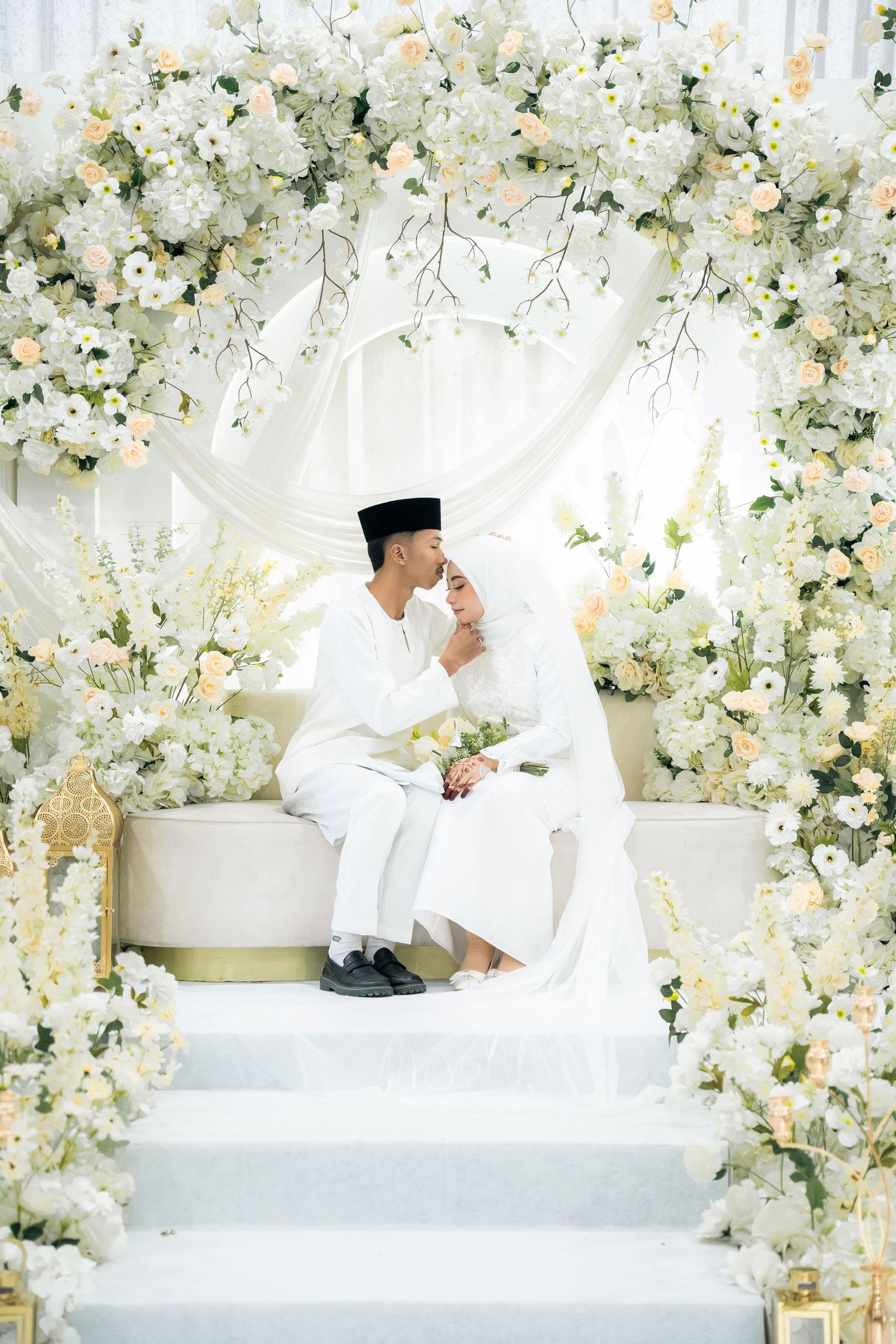 Muslim couple in white attire sitting on traditional wedding stage decorated with elegant white floral arrangements.