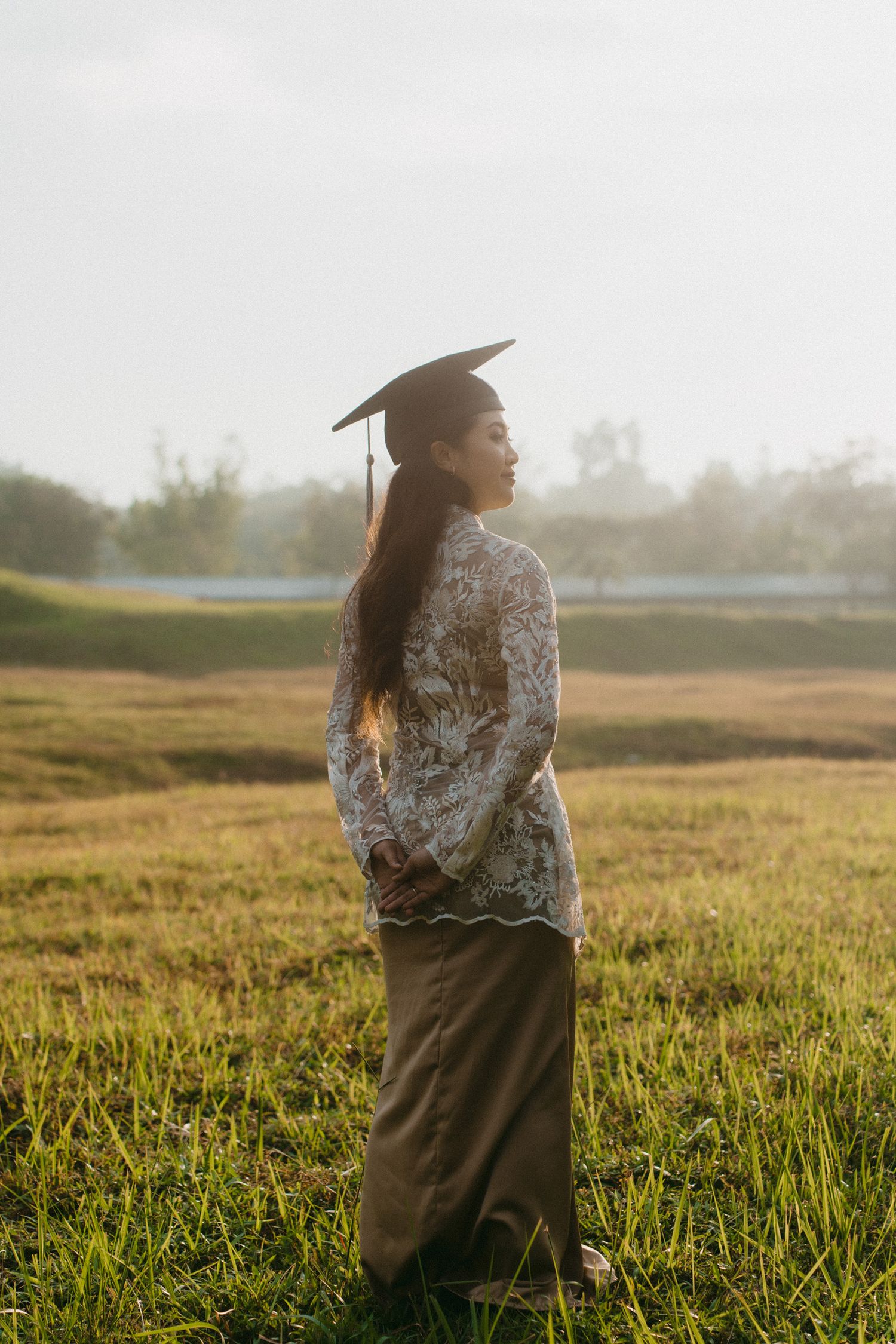Person standing in a sunlit field wearing graduation attire at golden hour.