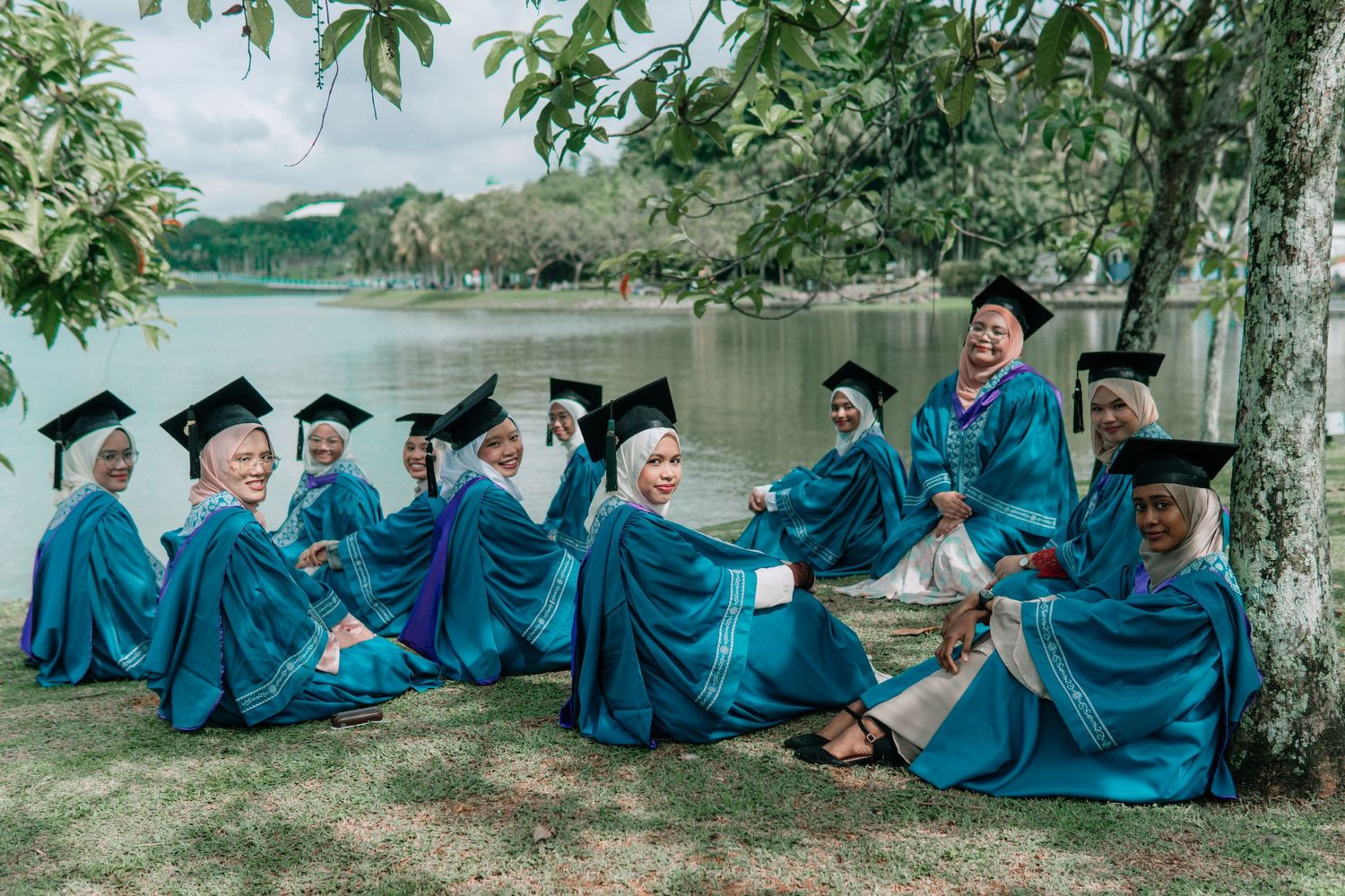 Graduates in blue gowns sitting on grass by a lake having a casual photoshoot.