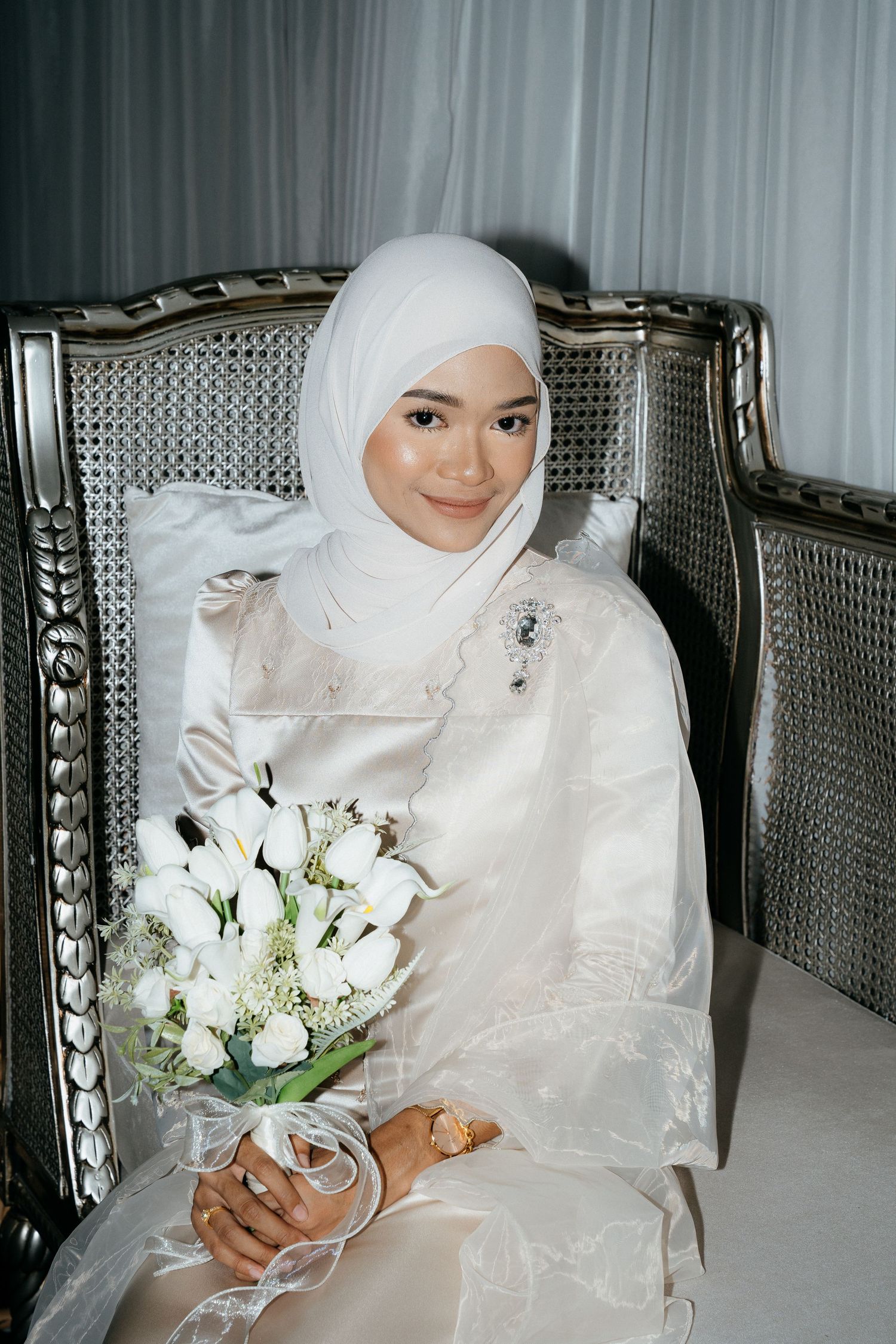 A bride in white hijab and dress holds a bouquet of white flowers while seated in an elegant vintage chair.