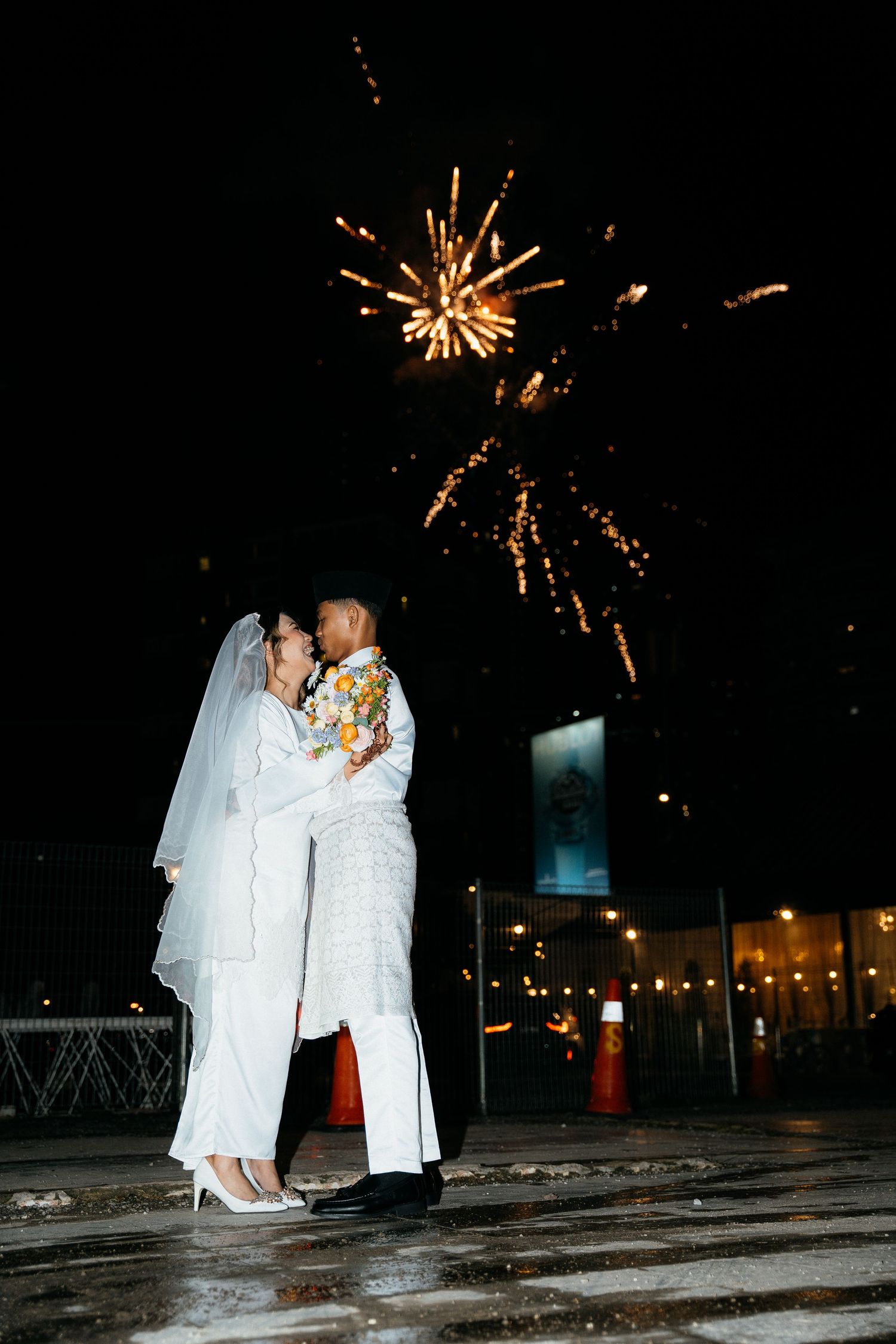 Two people in white attire share an intimate moment under sparkling fireworks on a dark evening.