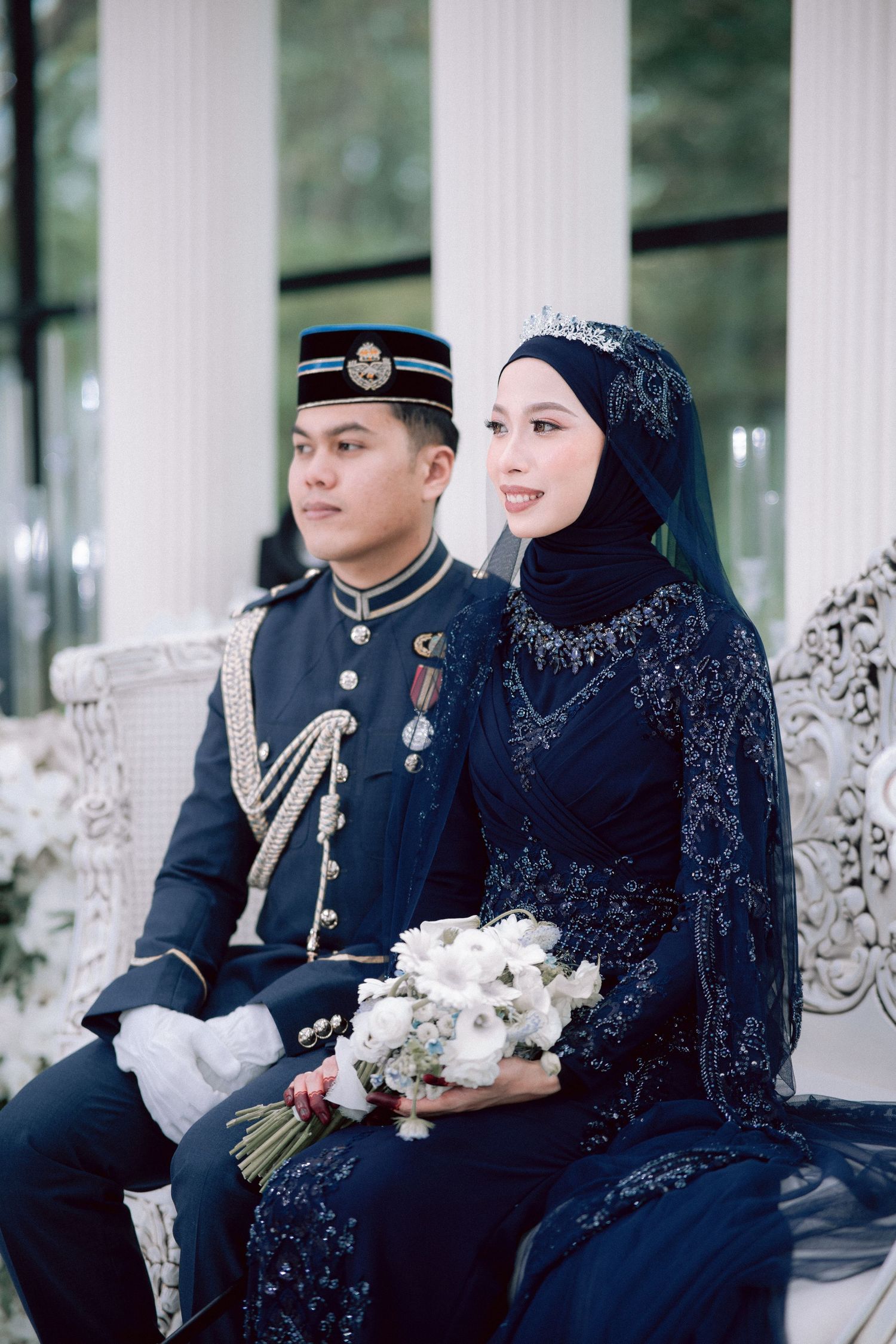 A couple in formal attire sits together outdoors, with the bride wearing a navy blue dress with ornate details.