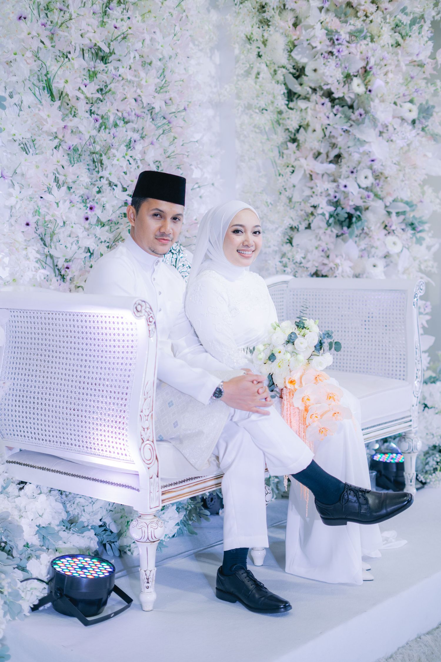 Muslim couple in traditional white wedding attire seated on elegant chairs against a floral backdrop.
