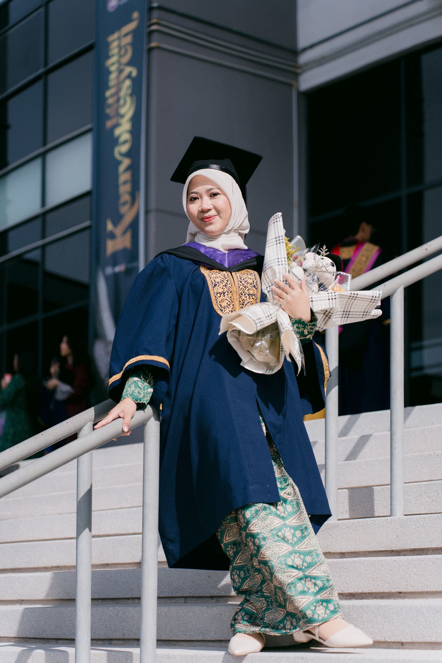 A graduate in a navy blue gown sits on stone steps while posing with their diploma in a celebratory sequence.