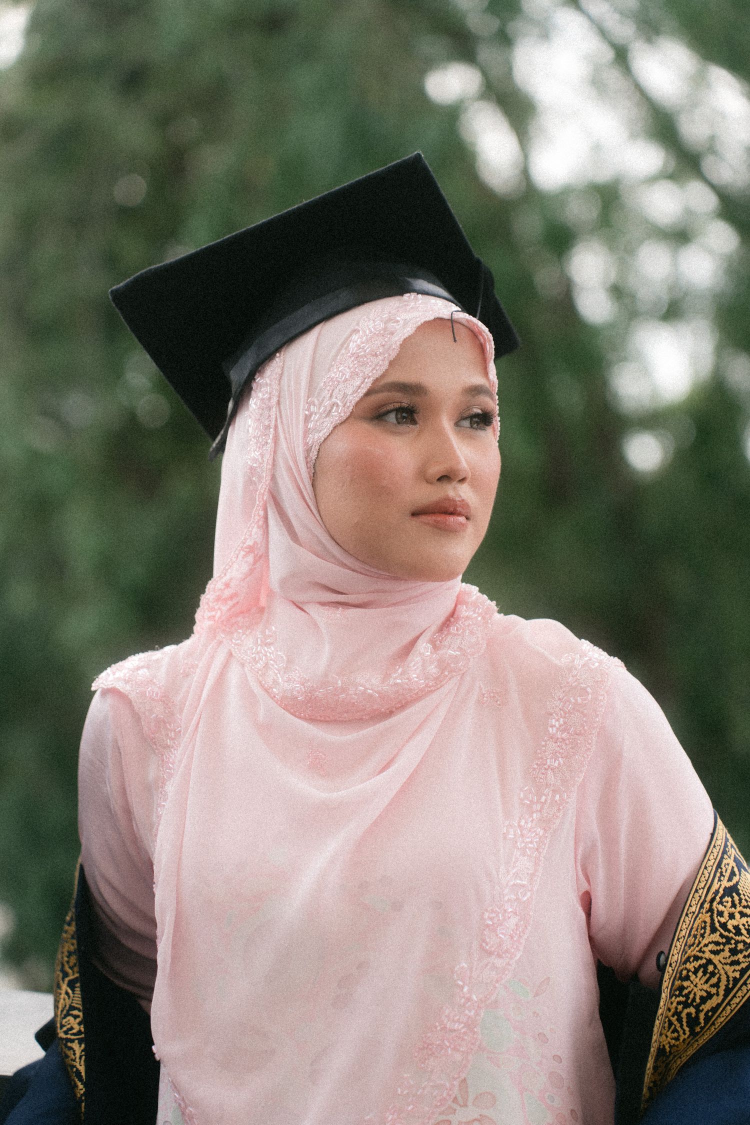A graduate wearing a pink hijab and black graduation cap looks thoughtfully into the distance during a ceremony.