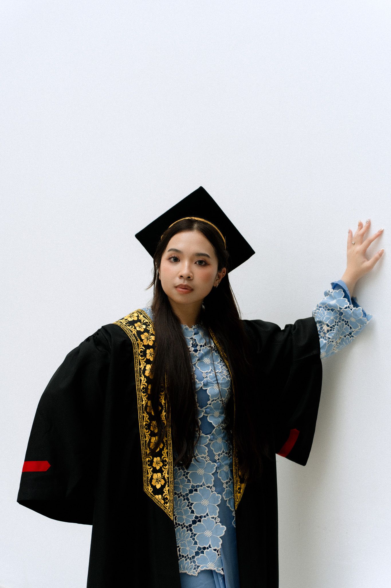 Young woman in black graduation gown with gold trim and mortarboard cap posing against a white wall.