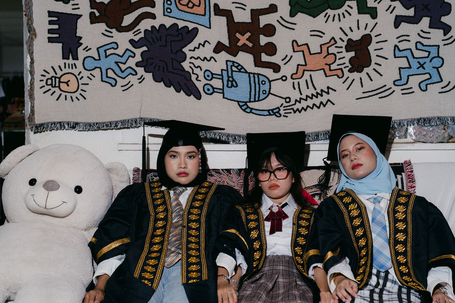 Three graduates in academic regalia pose with a large white teddy bear before a colorful Keith Haring-style mural.