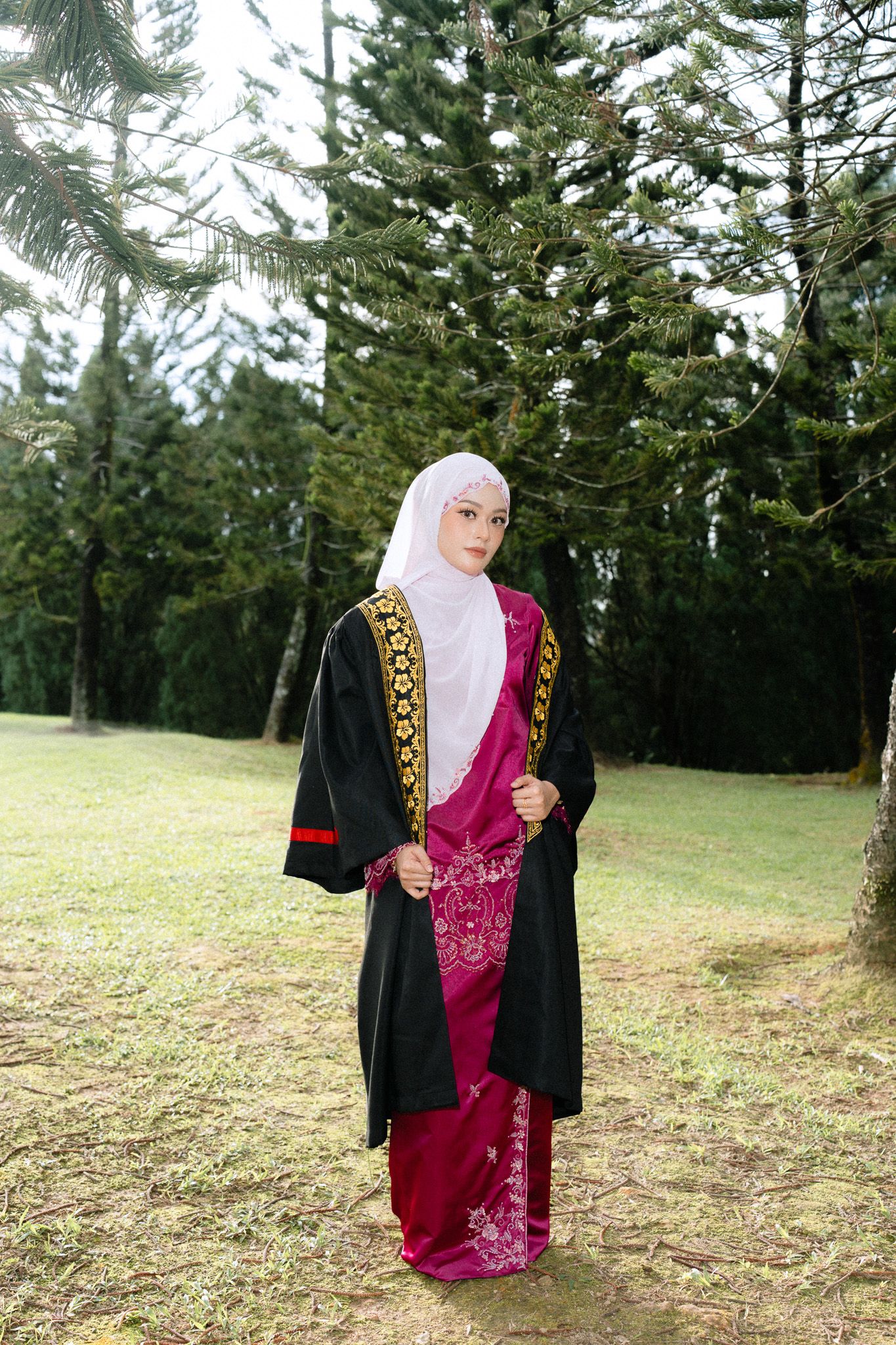 Muslim woman in black graduation gown and white hijab stands outdoors among pine trees wearing traditional pink attire.
