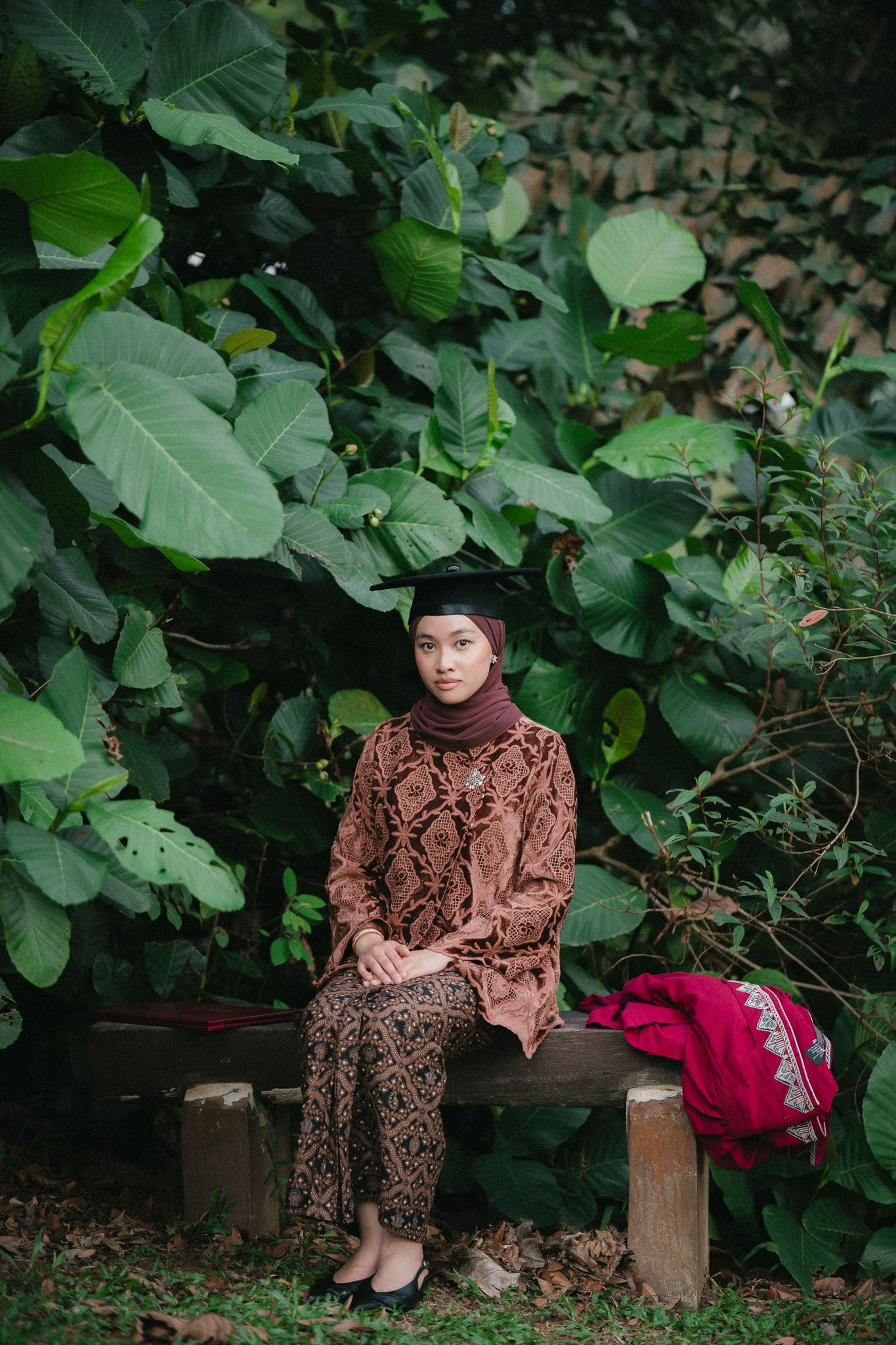 A young Muslim woman in traditional batik dress and graduation cap sits on a wooden bench surrounded by lush green tropical foliage.