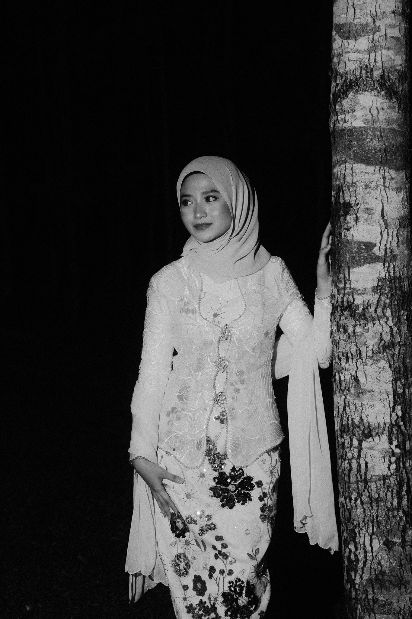 Black and white photo of a woman in hijab and floral kebaya dress posing beside a tree at night.