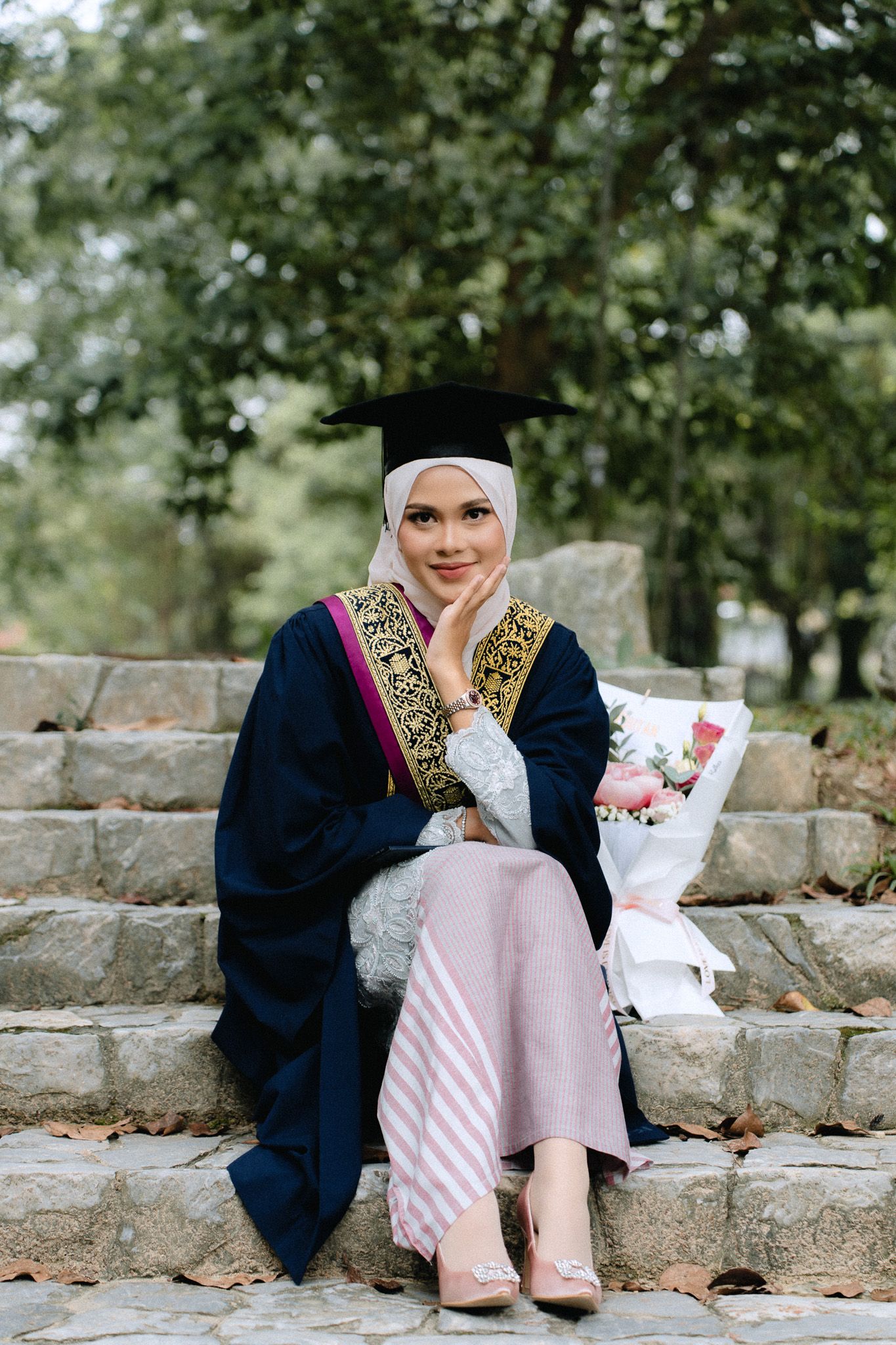 Muslim woman in graduation gown and hijab sits on steps holding flowers, smiling in an outdoor park setting.