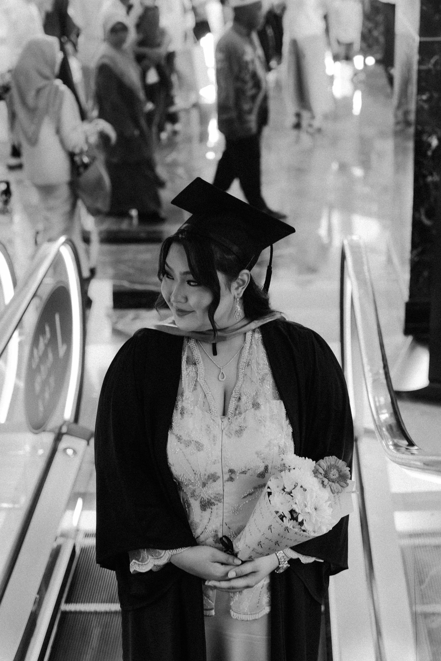 A graduate in black cap and gown holds a bouquet while standing on an escalator in a black and white photograph.
