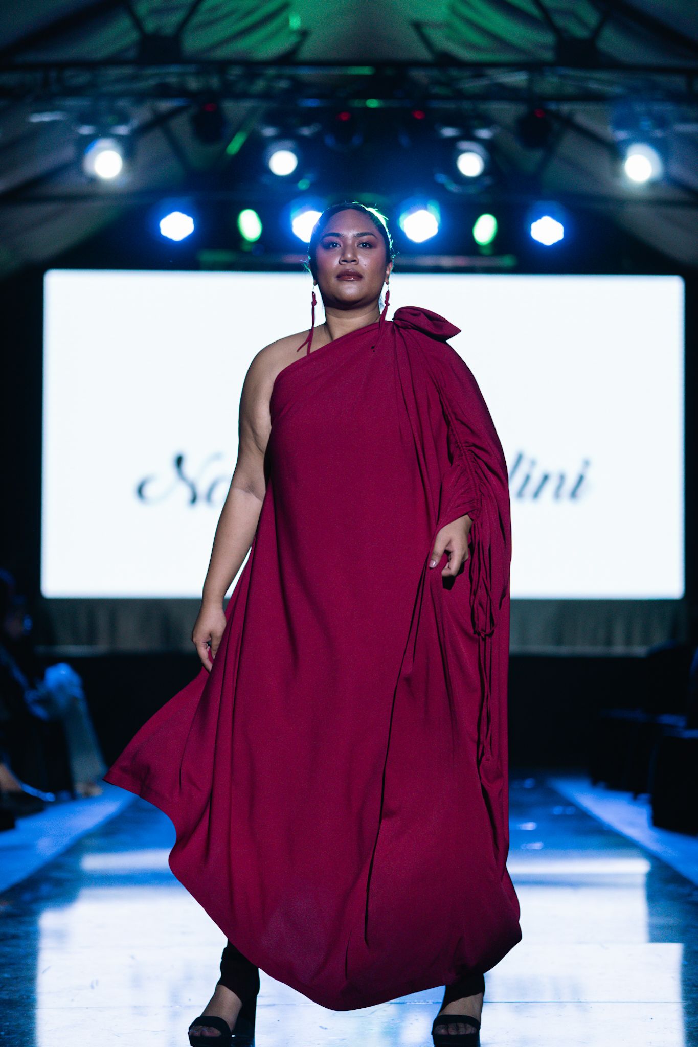 Model showcasing dramatic burgundy draped dress on illuminated fashion runway.