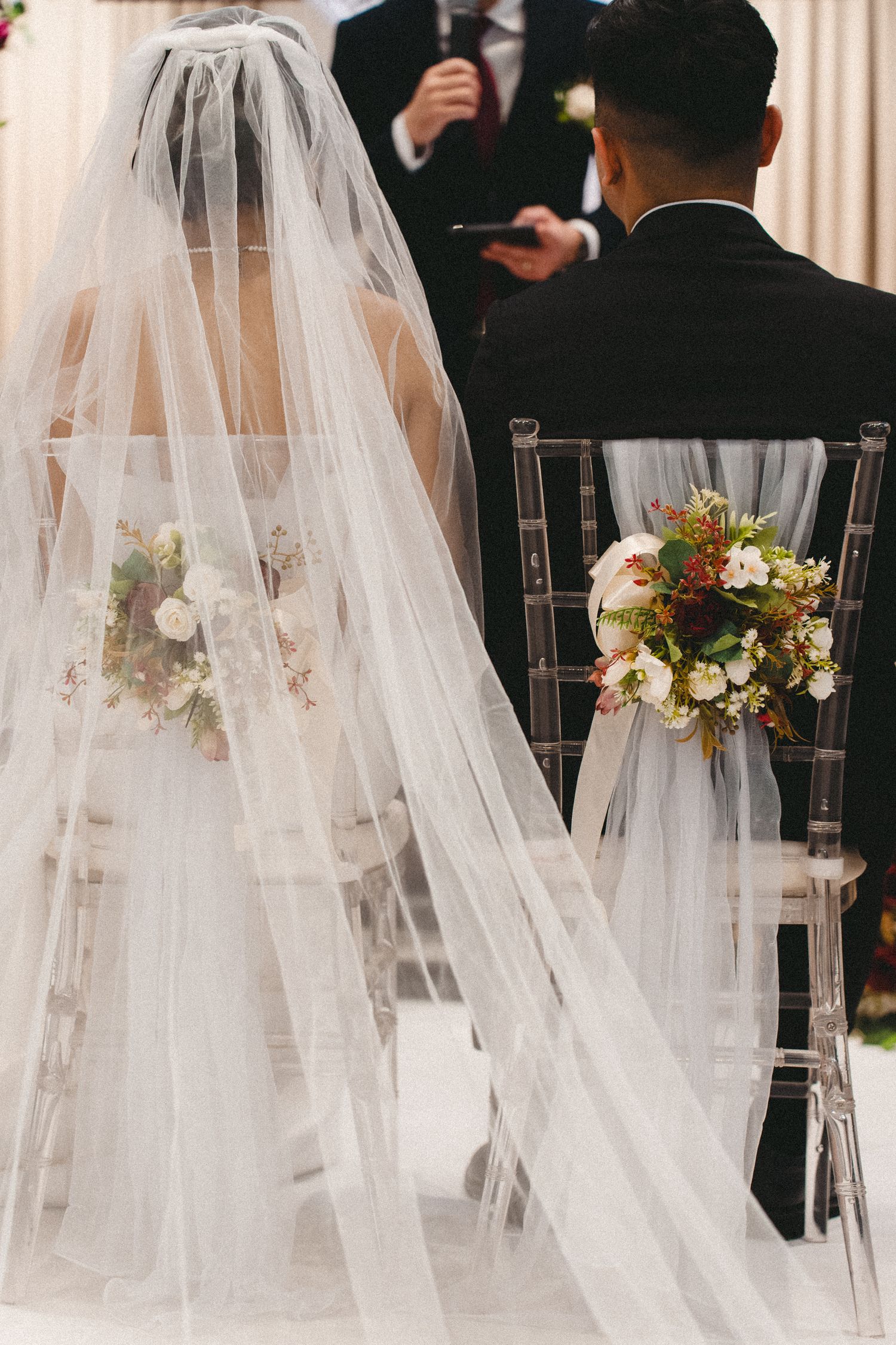 Elegant wedding scene with flowing veil and floral bouquet arrangements on clear chairs during ceremony.
