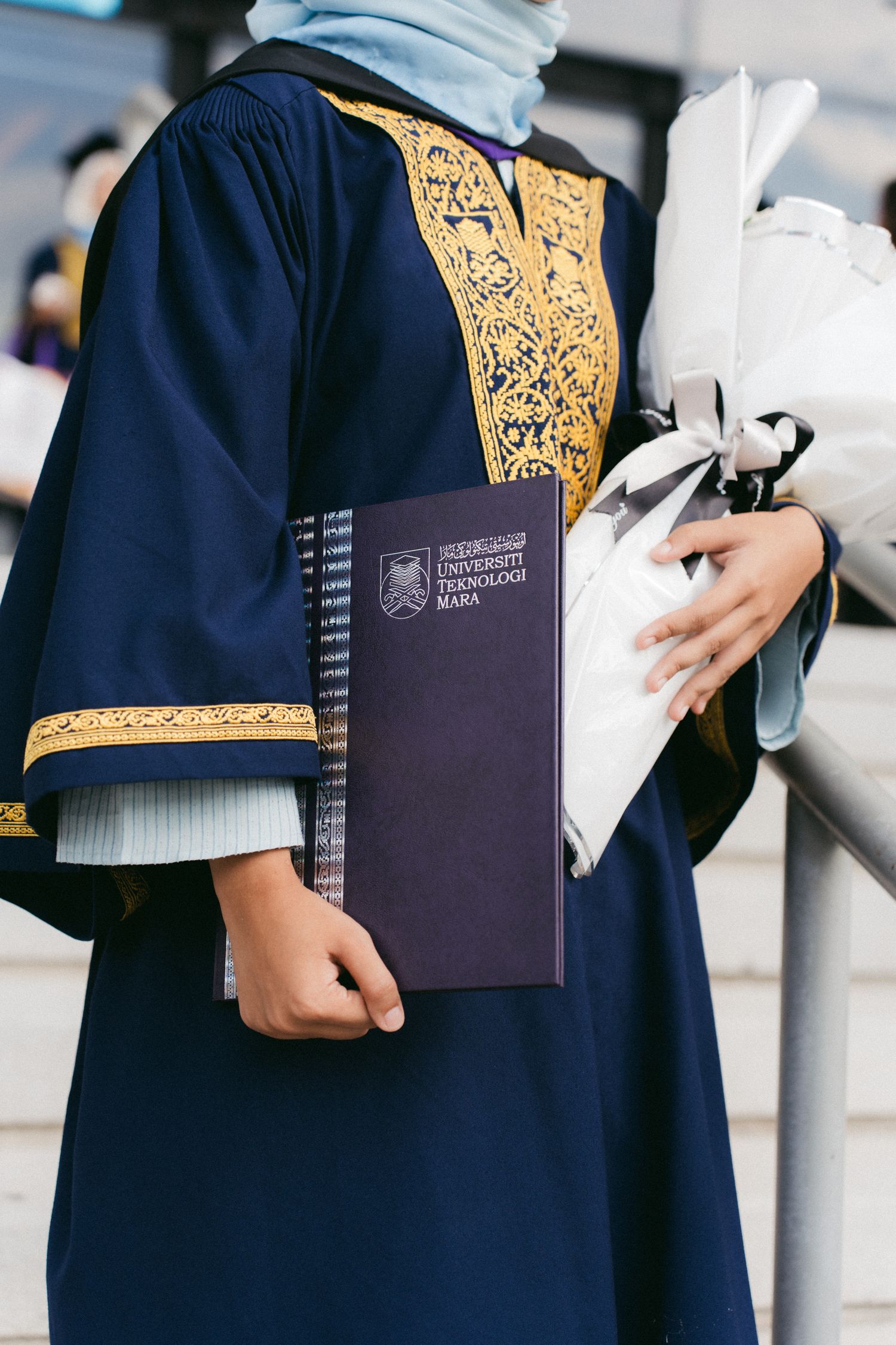 Graduate holding diploma in navy blue graduation gown with gold embellishments and light blue hijab.