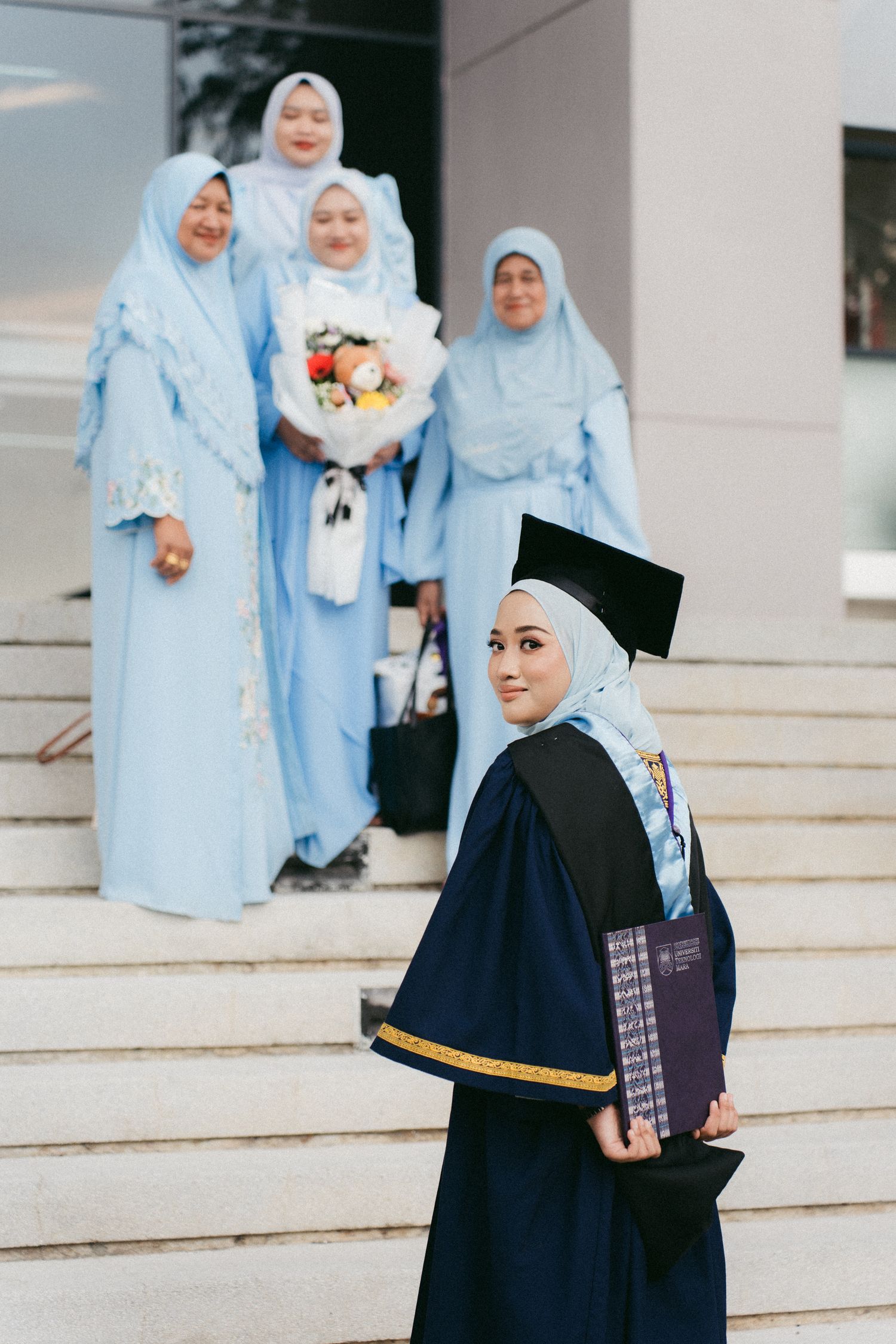 Graduation photo featuring women in light blue hijabs on stairs while a graduate in dark graduation robes holds diploma.
