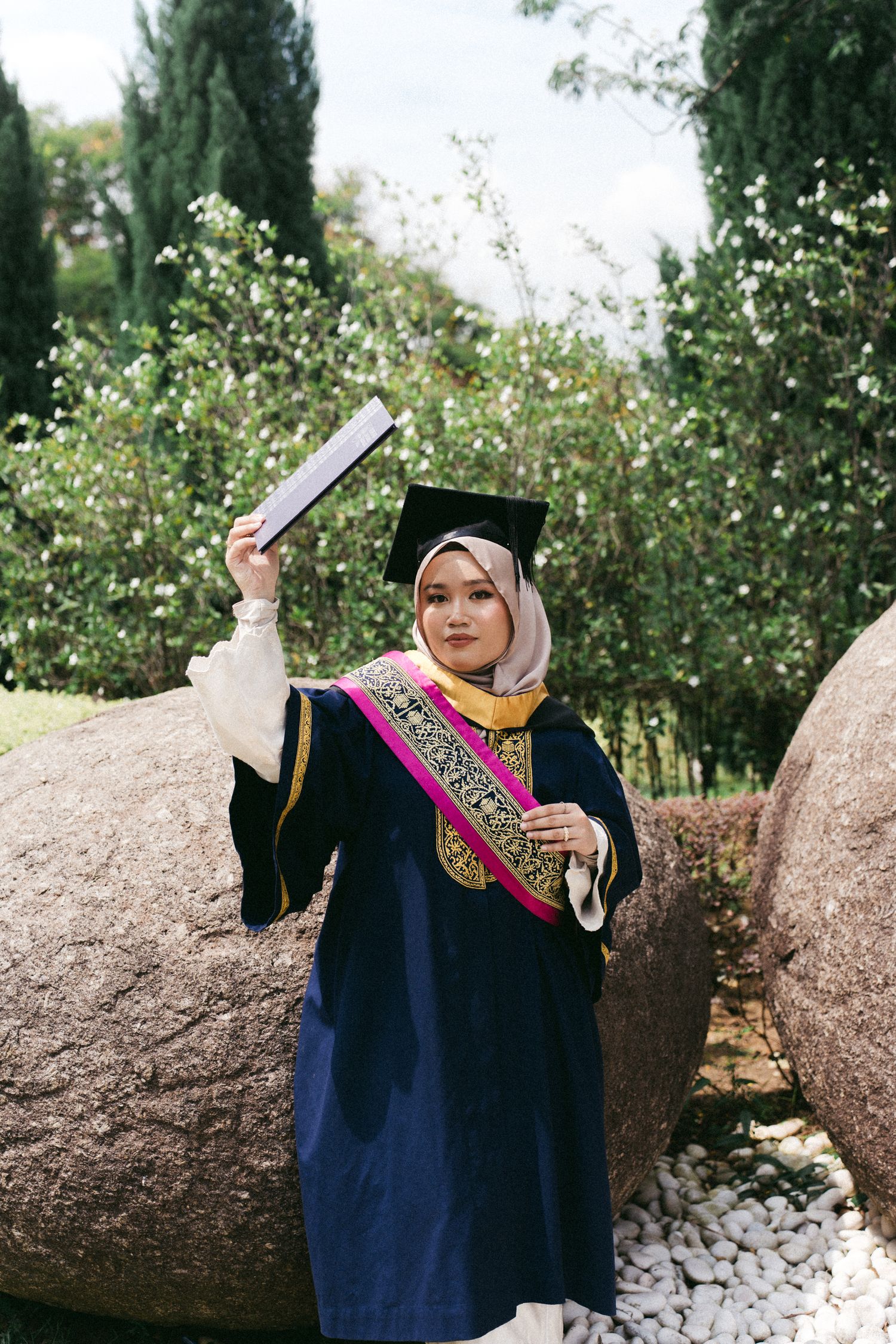 Graduate in navy graduation gown with decorative sash celebrates with diploma outdoors near garden rocks.