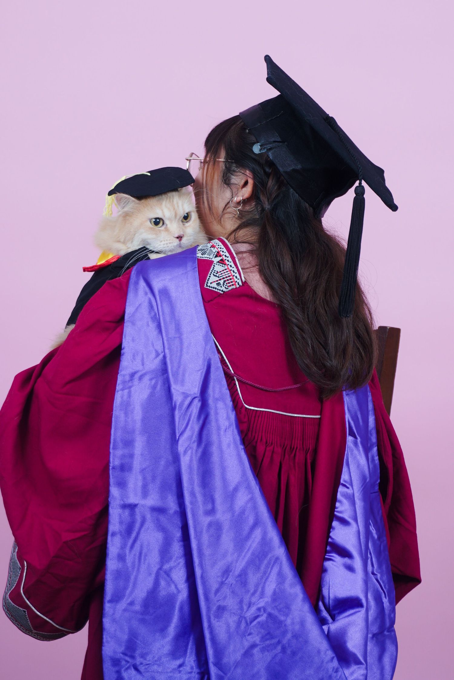 Graduate in red and purple academic gown with mortarboard holds a cat wearing a tiny graduation cap on pink background.
