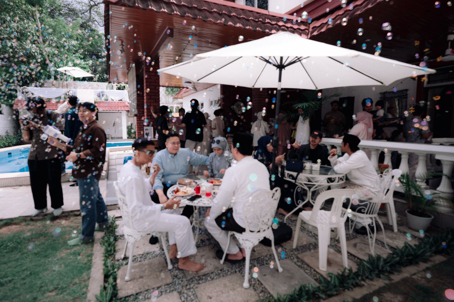 Group of people dining outdoors at a poolside restaurant under a white umbrella with bubbles floating in the air.