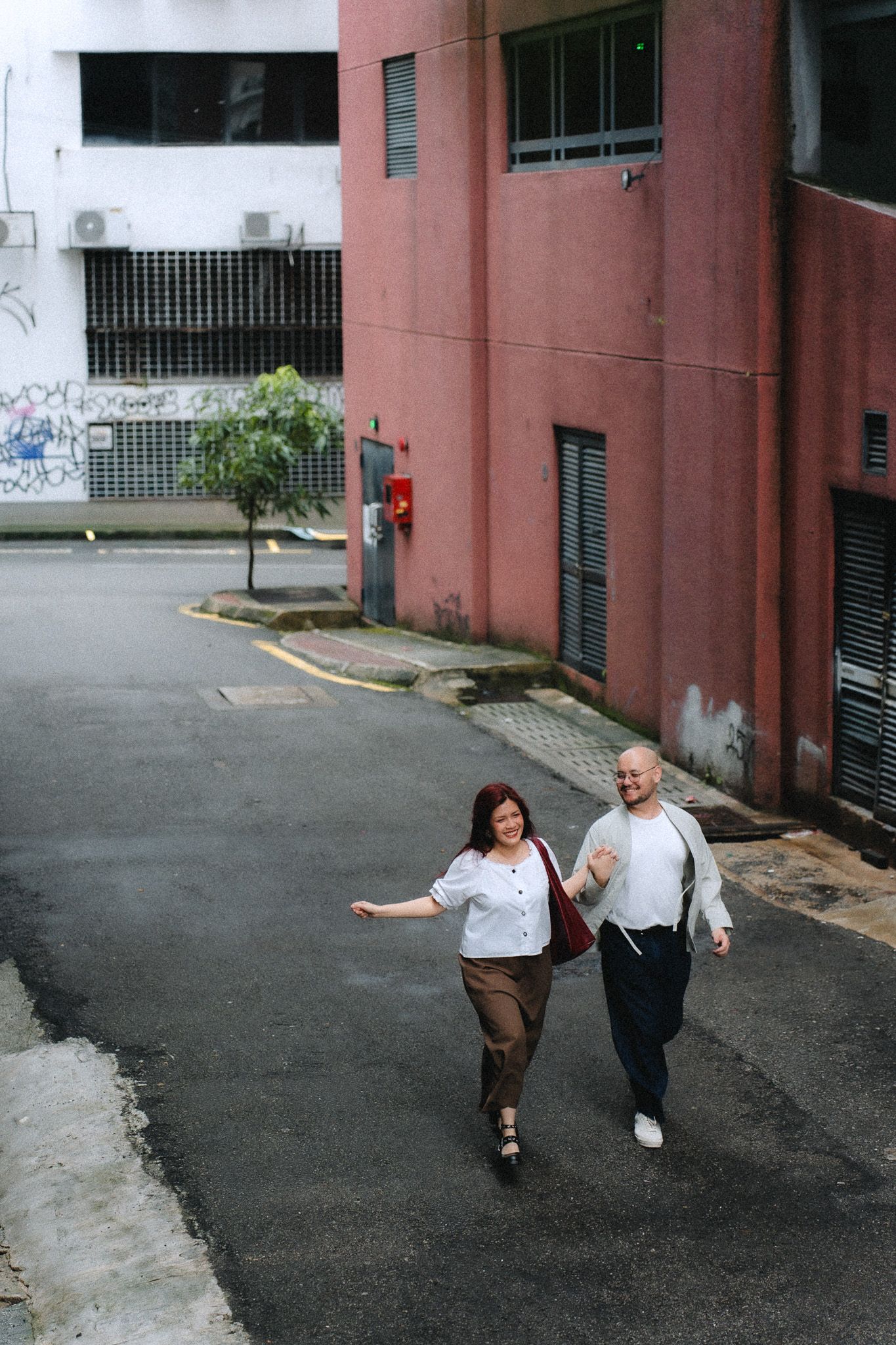 A cheerful couple walks together on an urban street beside a pink building, the woman carrying a red bag.