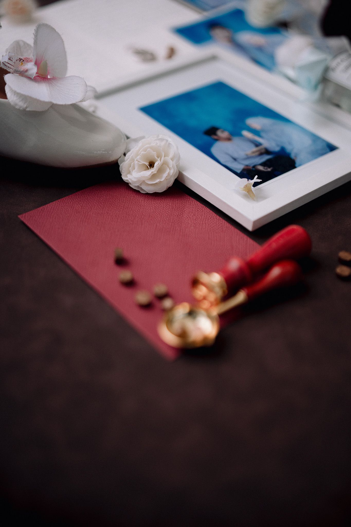 Elegant flat lay with white flowers, red envelope, wax seal stamp, coffee beans, and framed photo on dark surface.