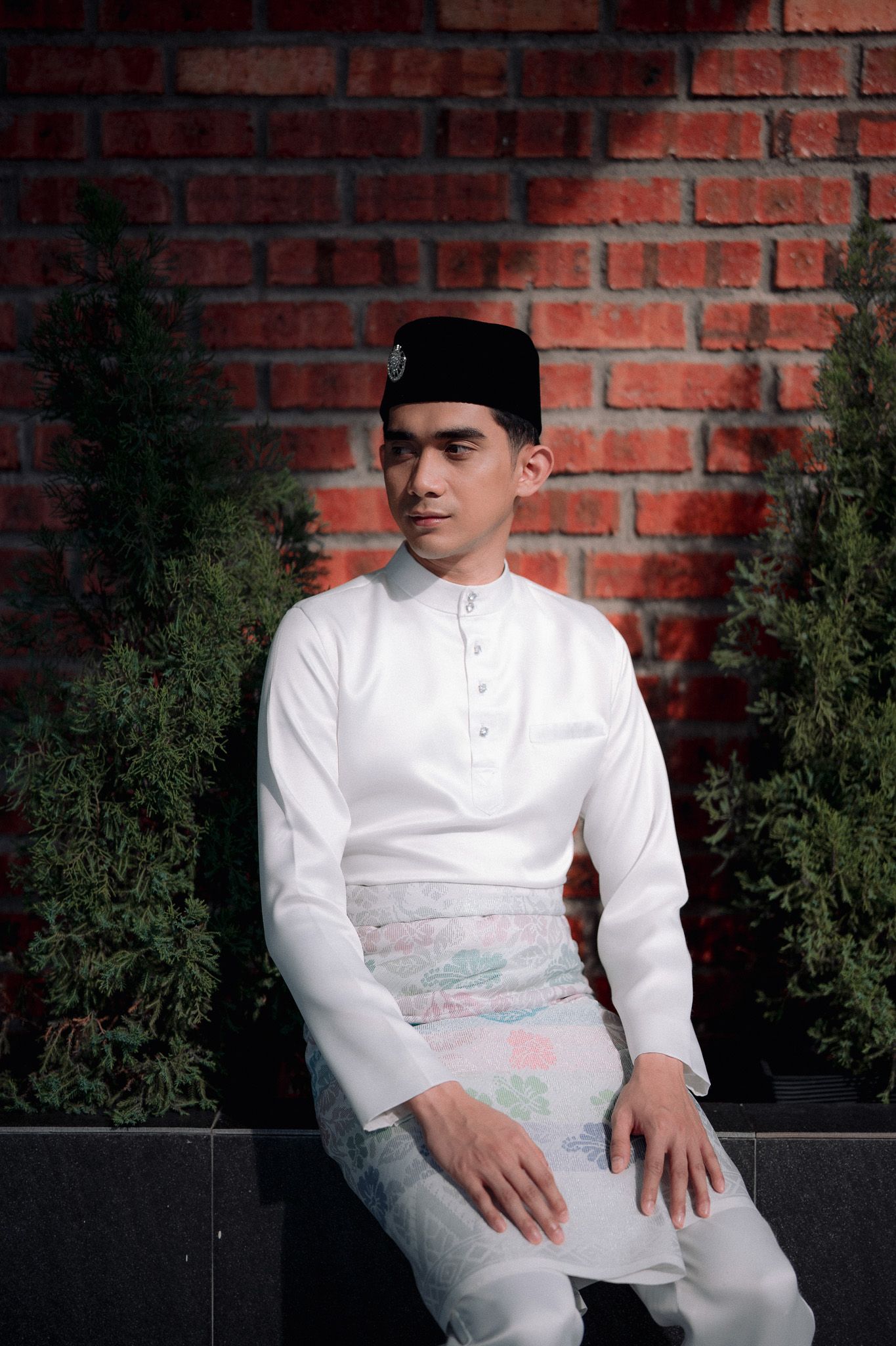 Young man in white Baju Melayu and songkok sitting against a red brick wall with green plants.