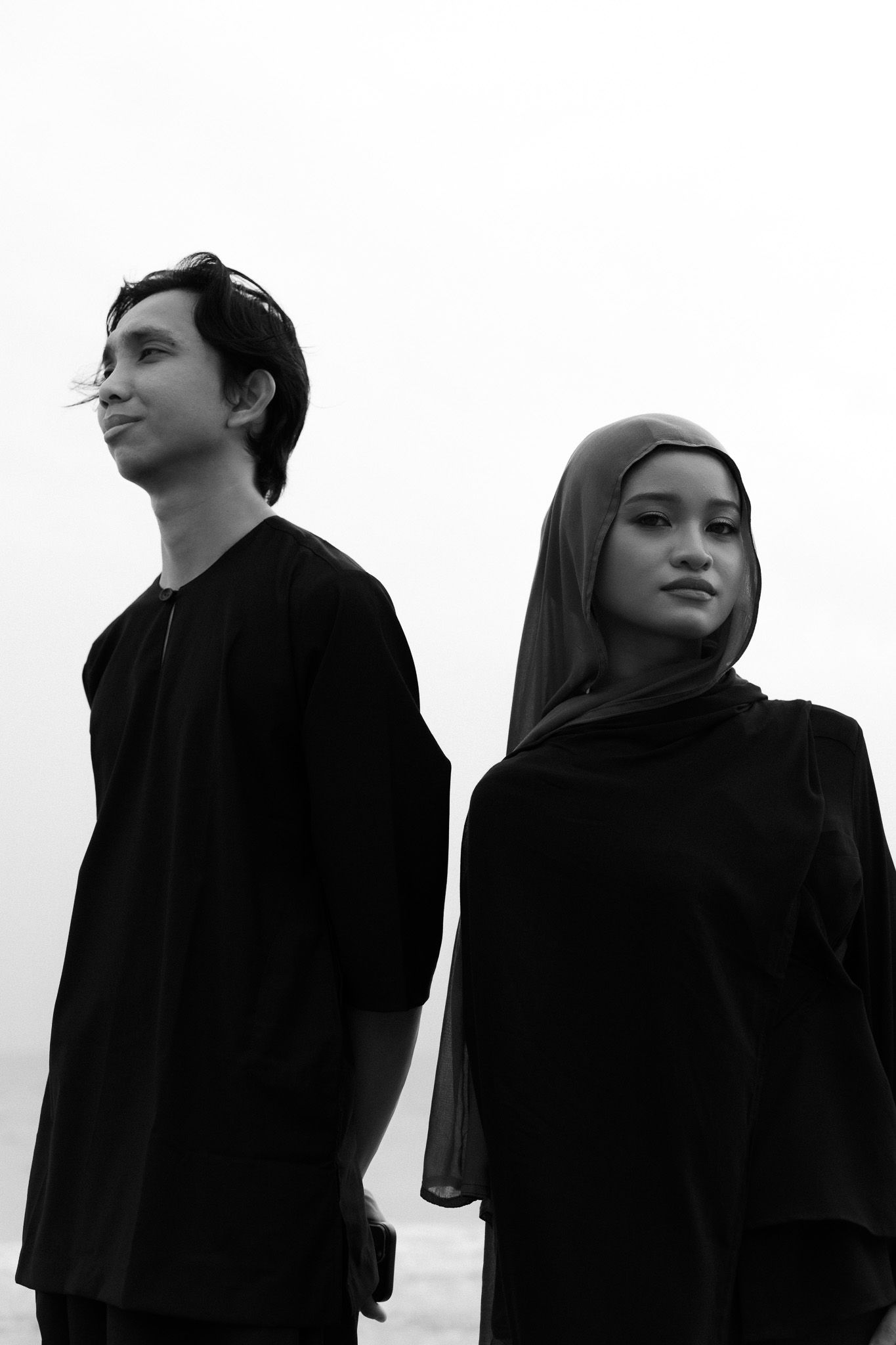 Young man and woman in black clothing and hijab posing together in black and white photo.