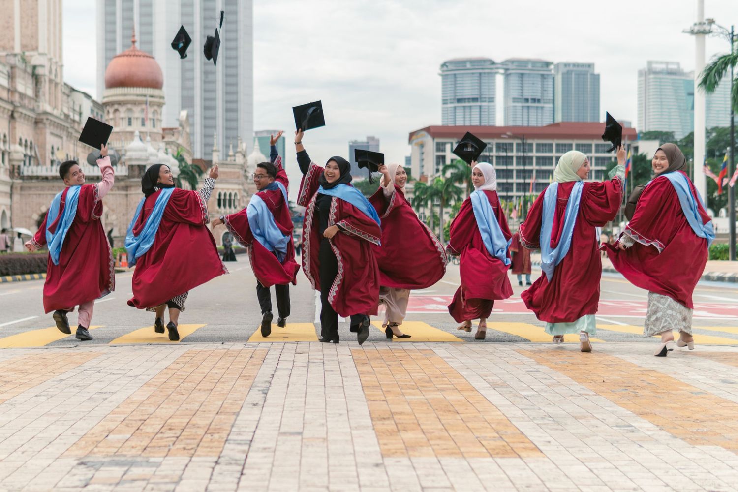 Graduates in red gowns jumping and celebrating on a city street with modern buildings in background.