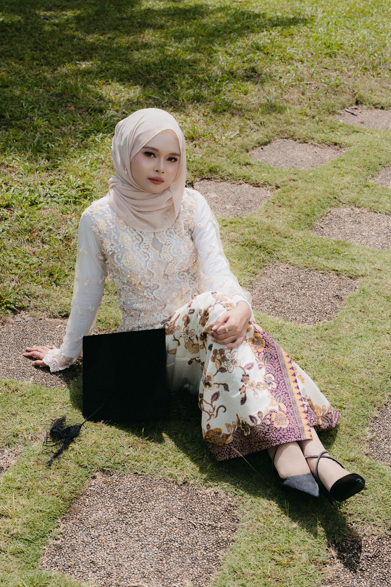 A young Muslim woman in white hijab and floral dress sits gracefully on grass steps with a laptop beside her.