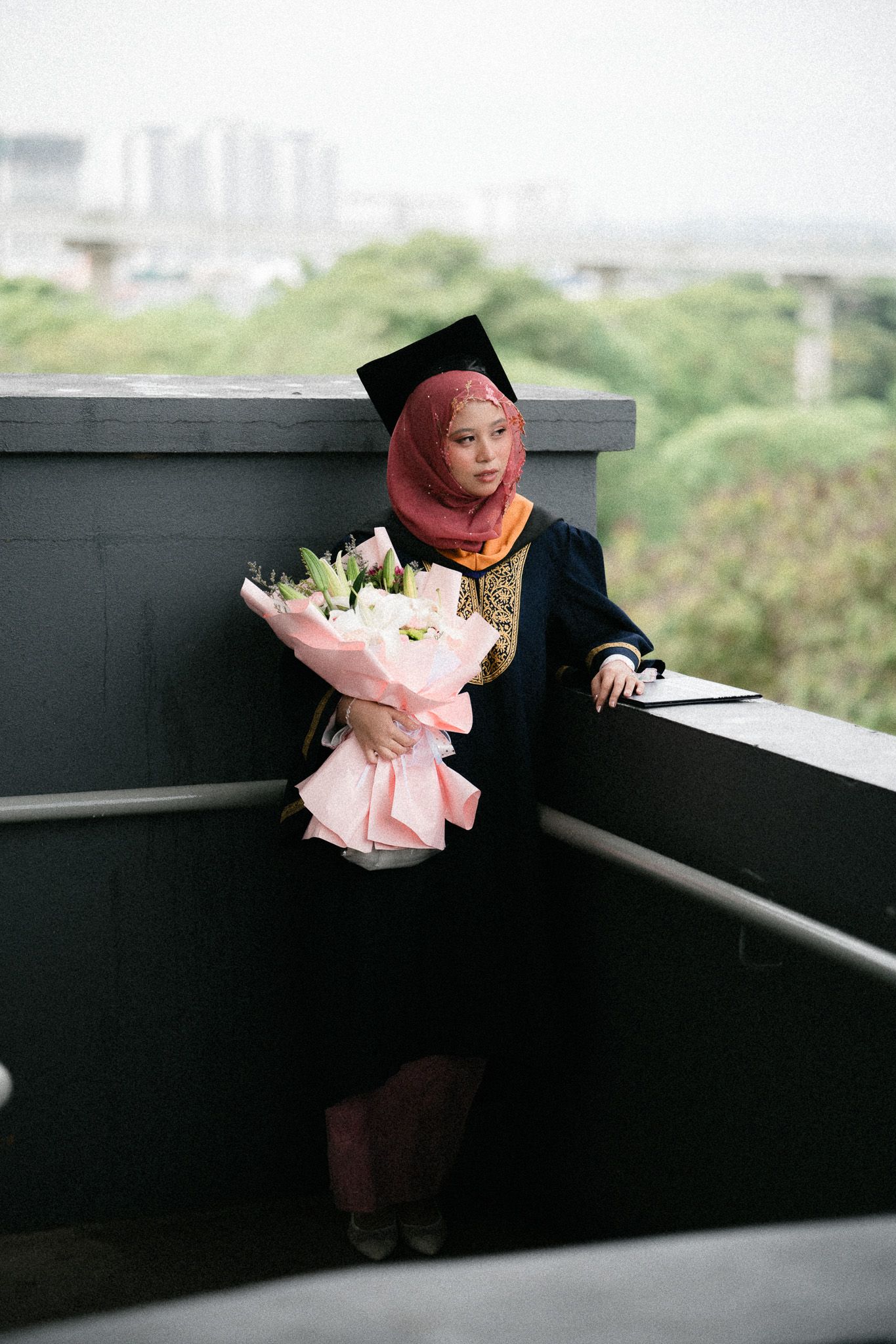 Graduate in black gown and cap holding pink flower bouquet on balcony wearing red hijab.