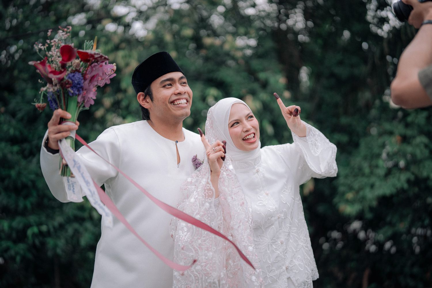 Happy Malaysian couple in traditional white wedding attire celebrating outdoors, groom holding colorful bouquet, bride gesturing joyfully.