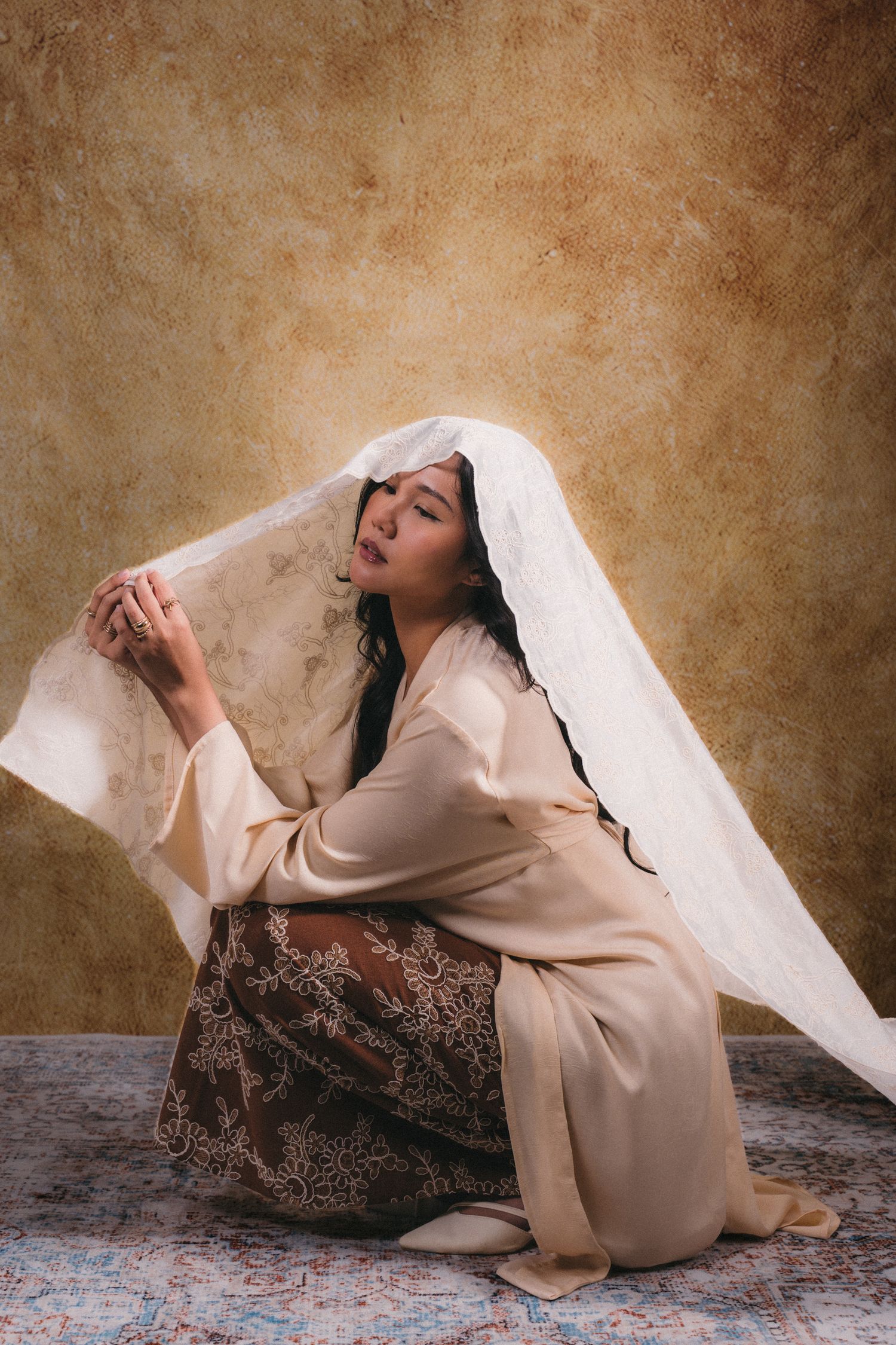 An artistic portrait of a person in beige clothing with lace veil against brown textured backdrop.
