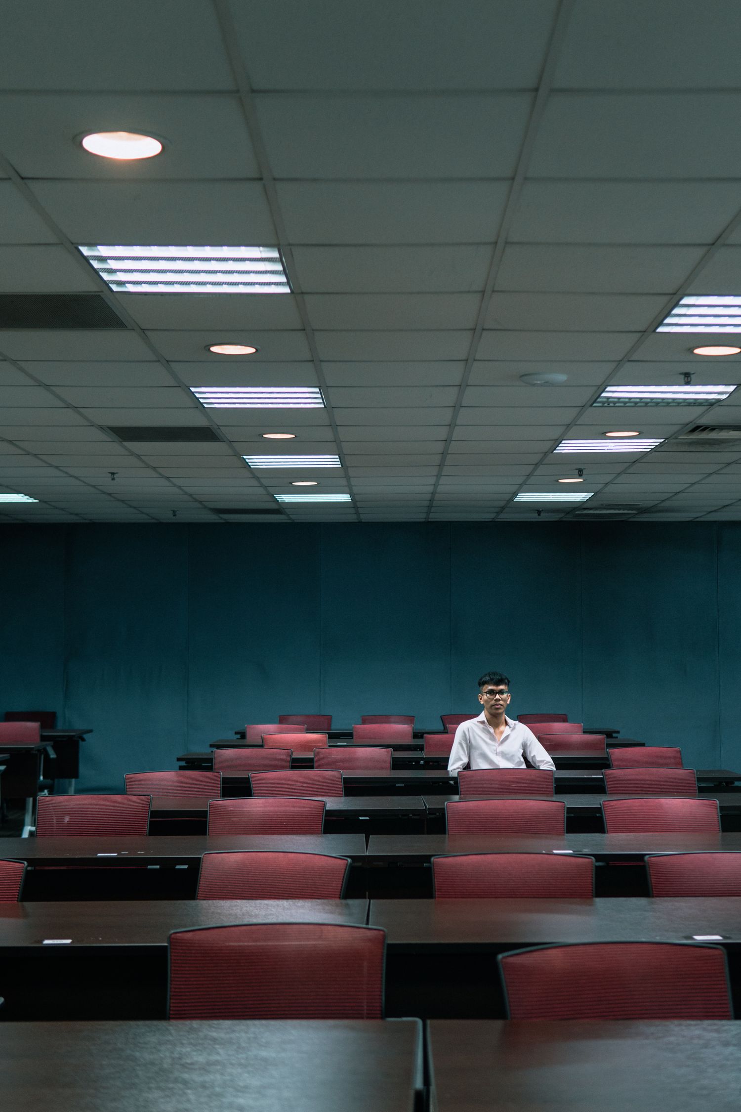 Empty lecture hall with red seats shows a solitary figure seated in the middle of rows of chairs.