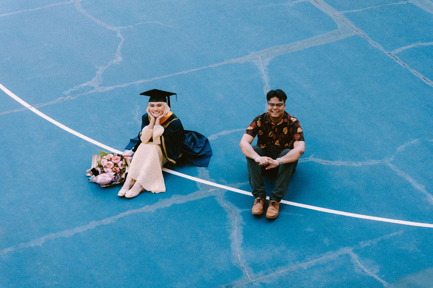 Two people sitting on a blue basketball court with white lines during graduation.