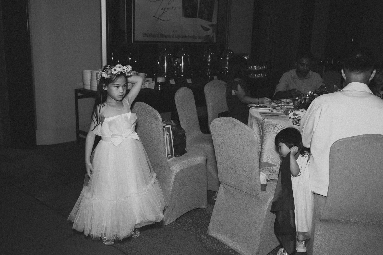 A young attendee in a white dress and flower crown stands near dining tables at a formal event.