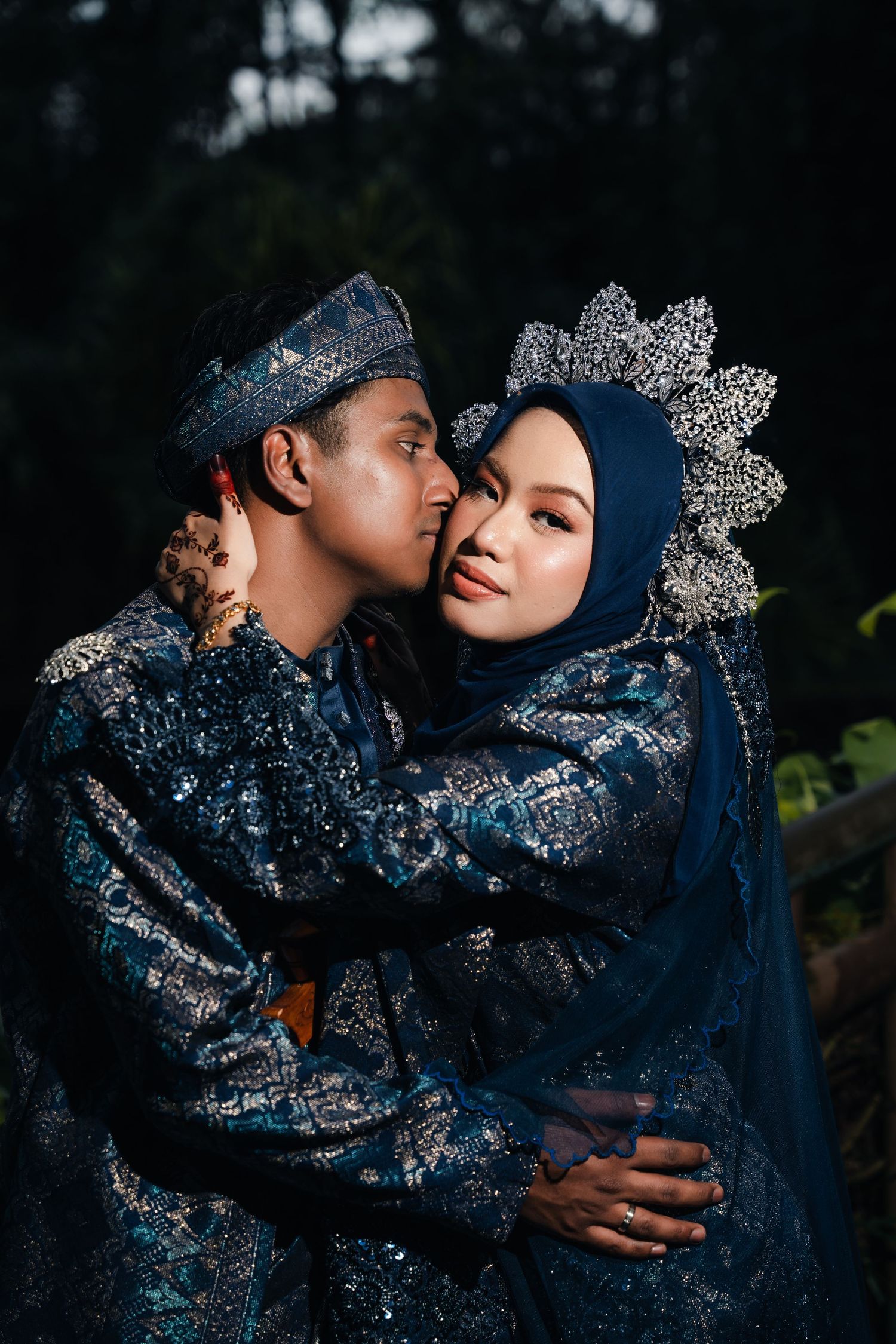 Traditional Malay wedding couple wearing matching ornate navy blue traditional attire with silver embellishments embracing.