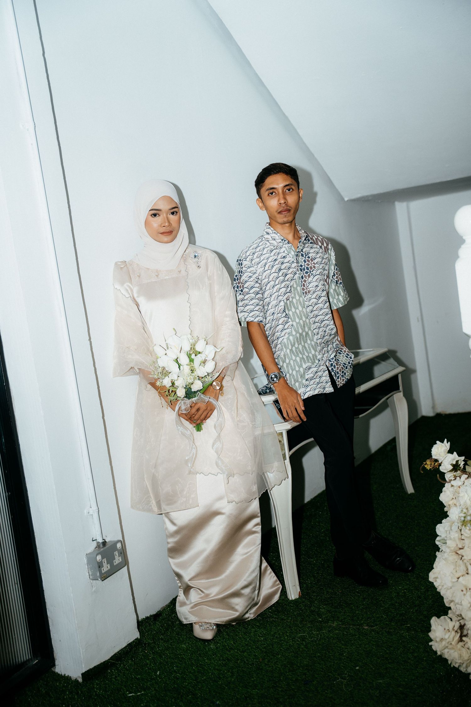 A wedding couple in traditional ceremony attire pose in a white interior space with floral decorations.