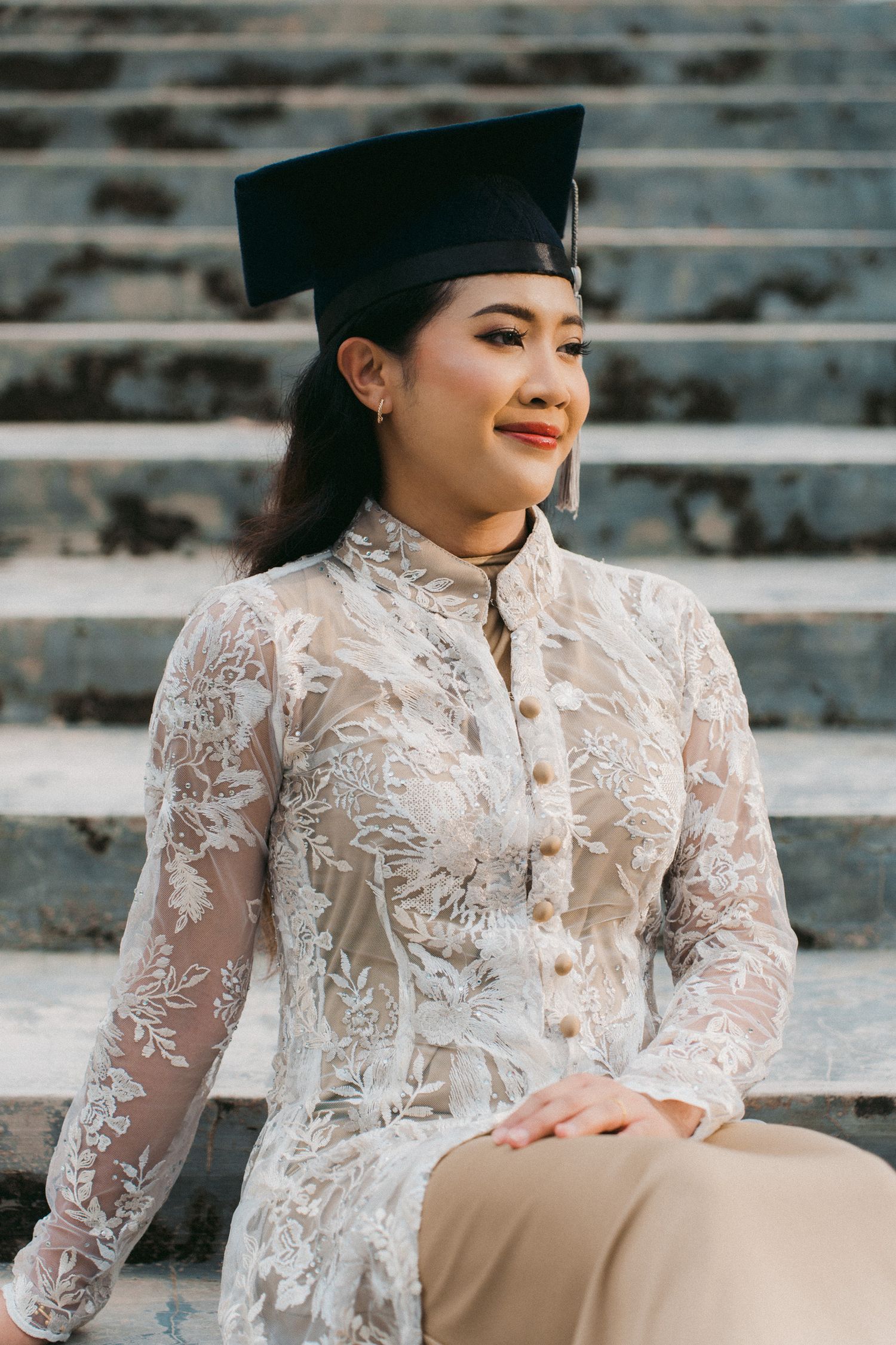 Graduate wearing elegant white lace blouse and graduation cap sits on concrete steps for portrait session.